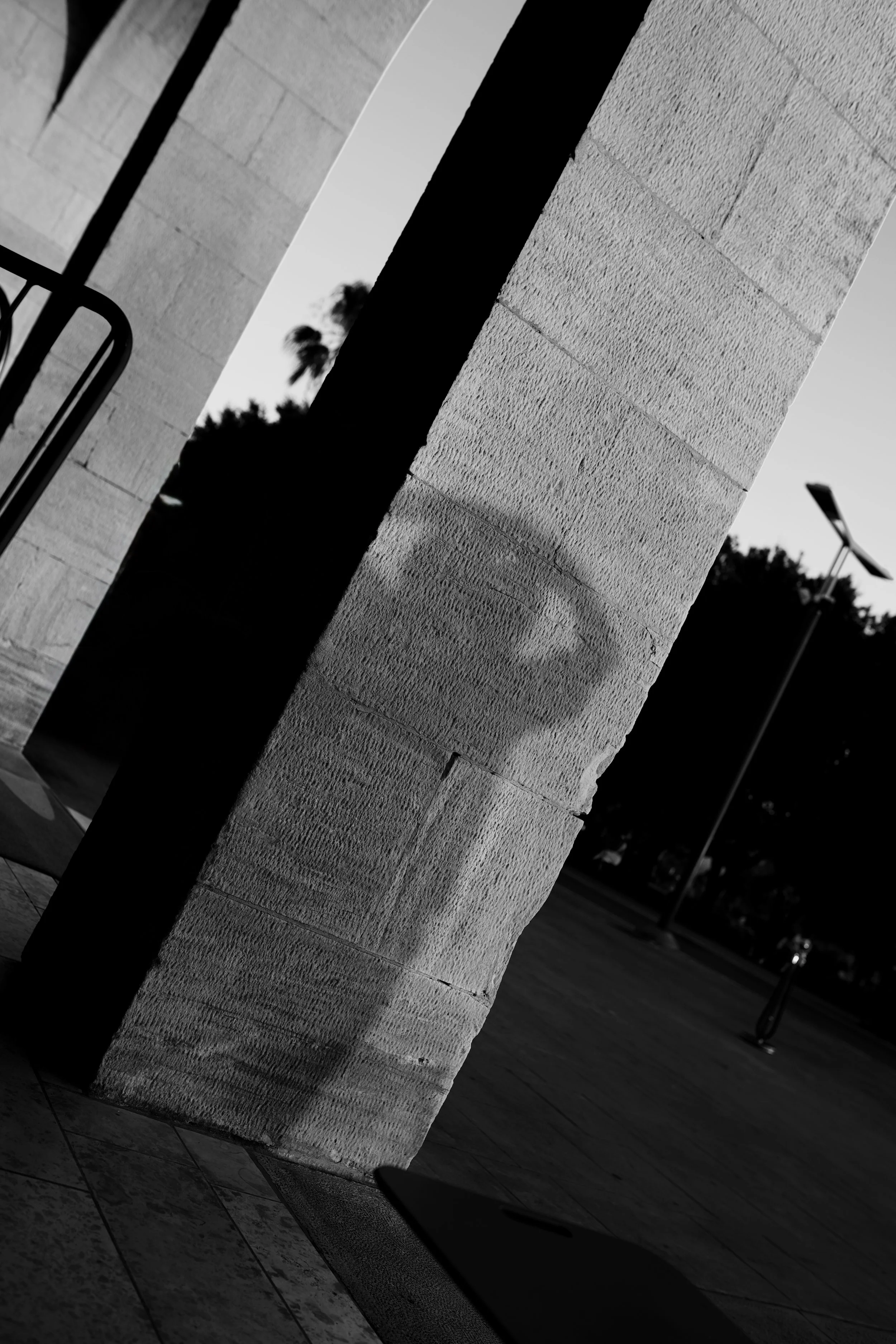 Black and white photo of a textured stone wall with an archway and a metal railing, likely part of a building's exterior, with trees and a street visible in the background.