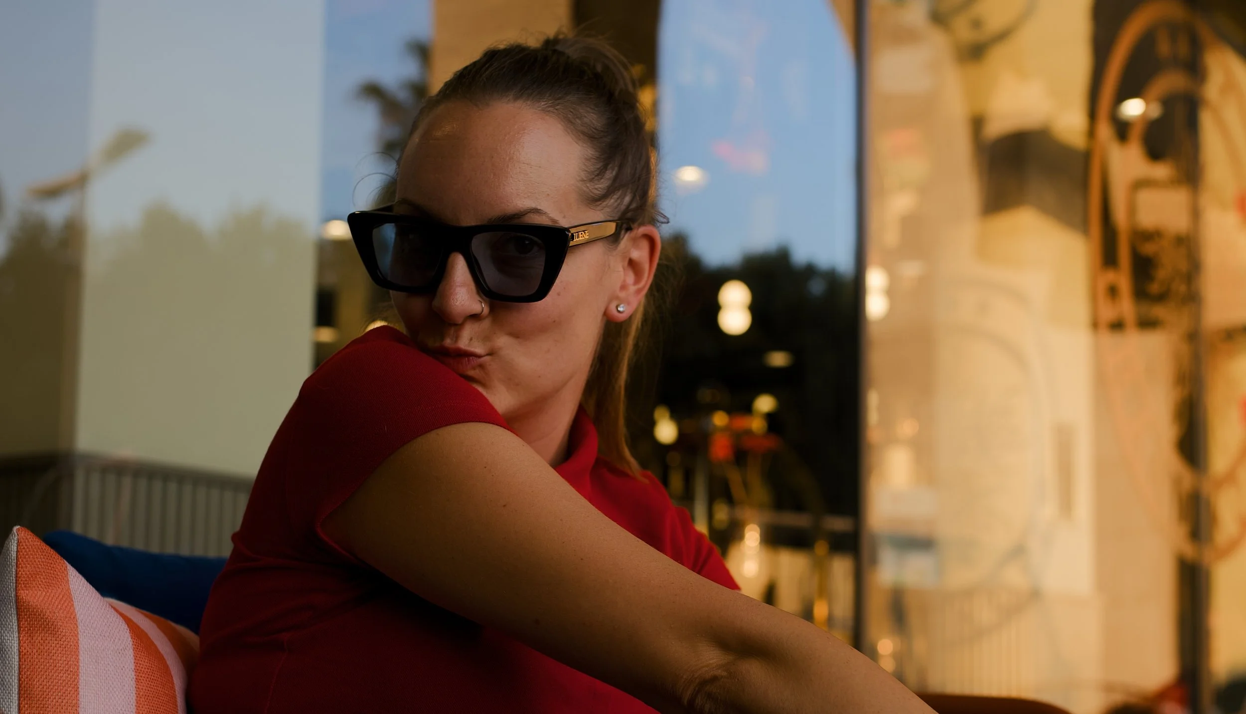 A woman with glasses and a nose piercing in a red shirt, sitting outside during sunset with warm lighting, looking over her shoulder.