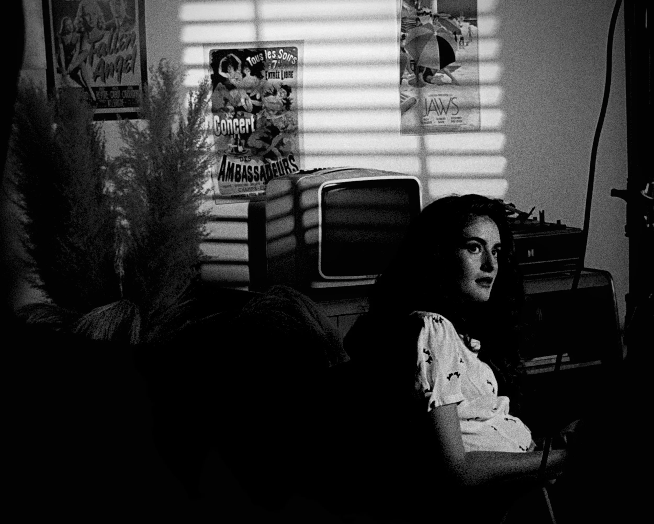 Black and white photo of a woman sitting in a room with posters on the wall, a TV on a table, and shadows of window blinds on the wall.