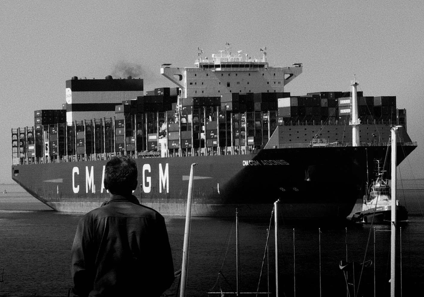 Person standing on dock observing a large cargo ship with containers, labeled 'CMA CGM', in the water with a tugboat nearby.