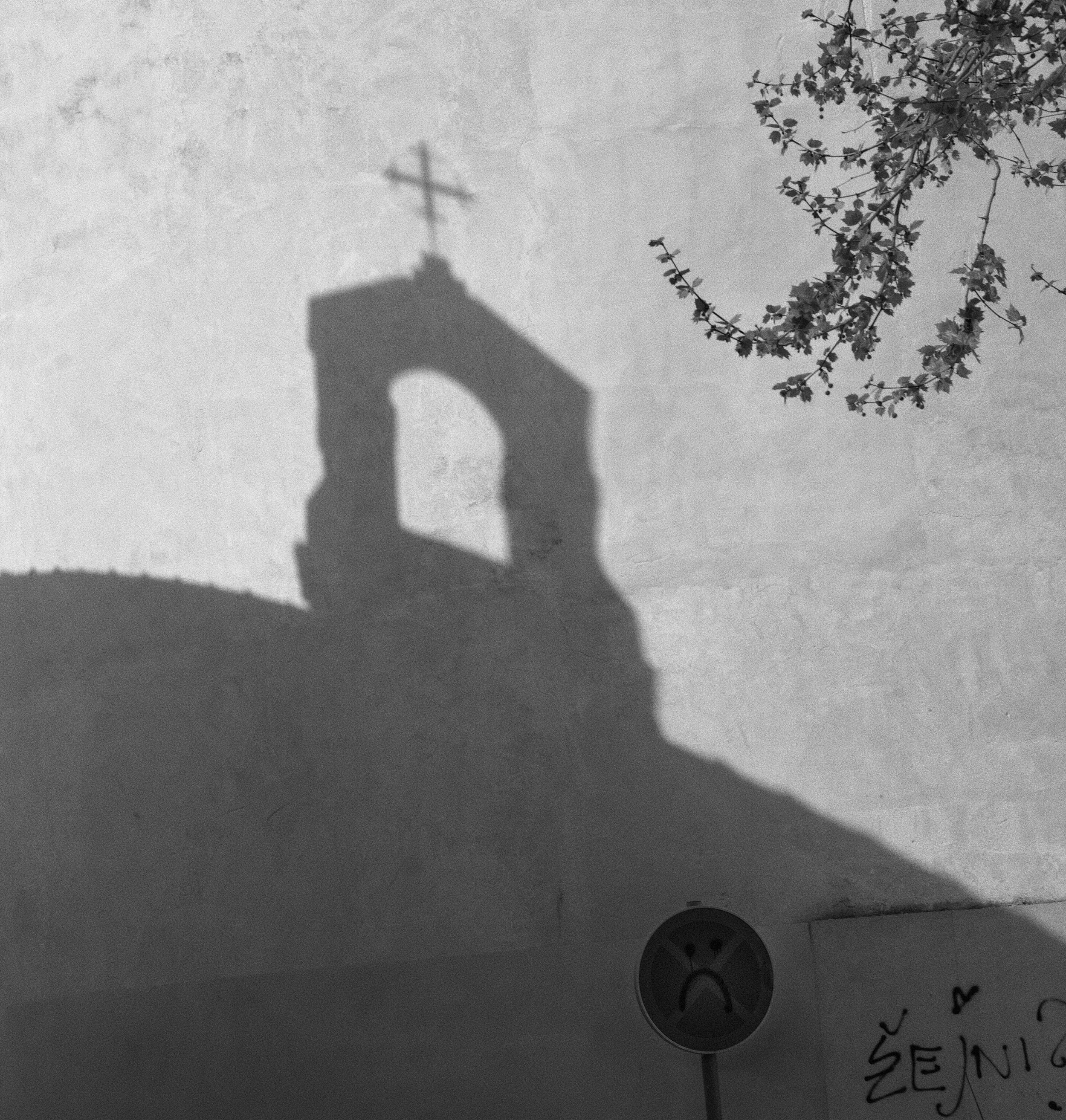 Black-and-white photo of a wall with a shadow of a cross that is cast from an angled angle. There is a branch with leaves hanging down from the top right corner. In the bottom right corner, there is a paper with handwritten text that reads 'SENT' and