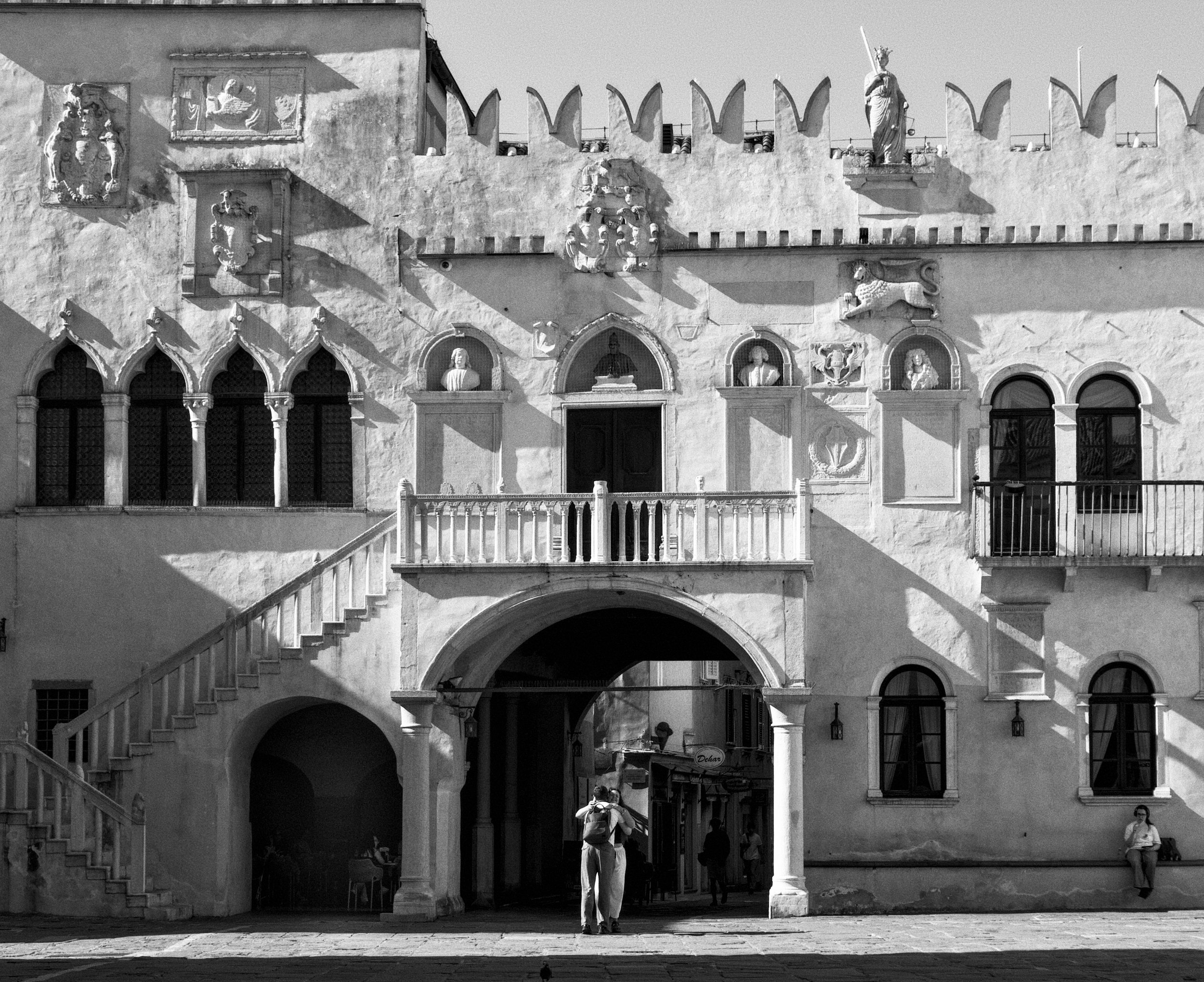 A black and white photo of an old building with ornate architectural decorations, arched windows, and statues on the facade. There are some people walking and standing in front of the building, including a person with a backpack taking a photo. The b