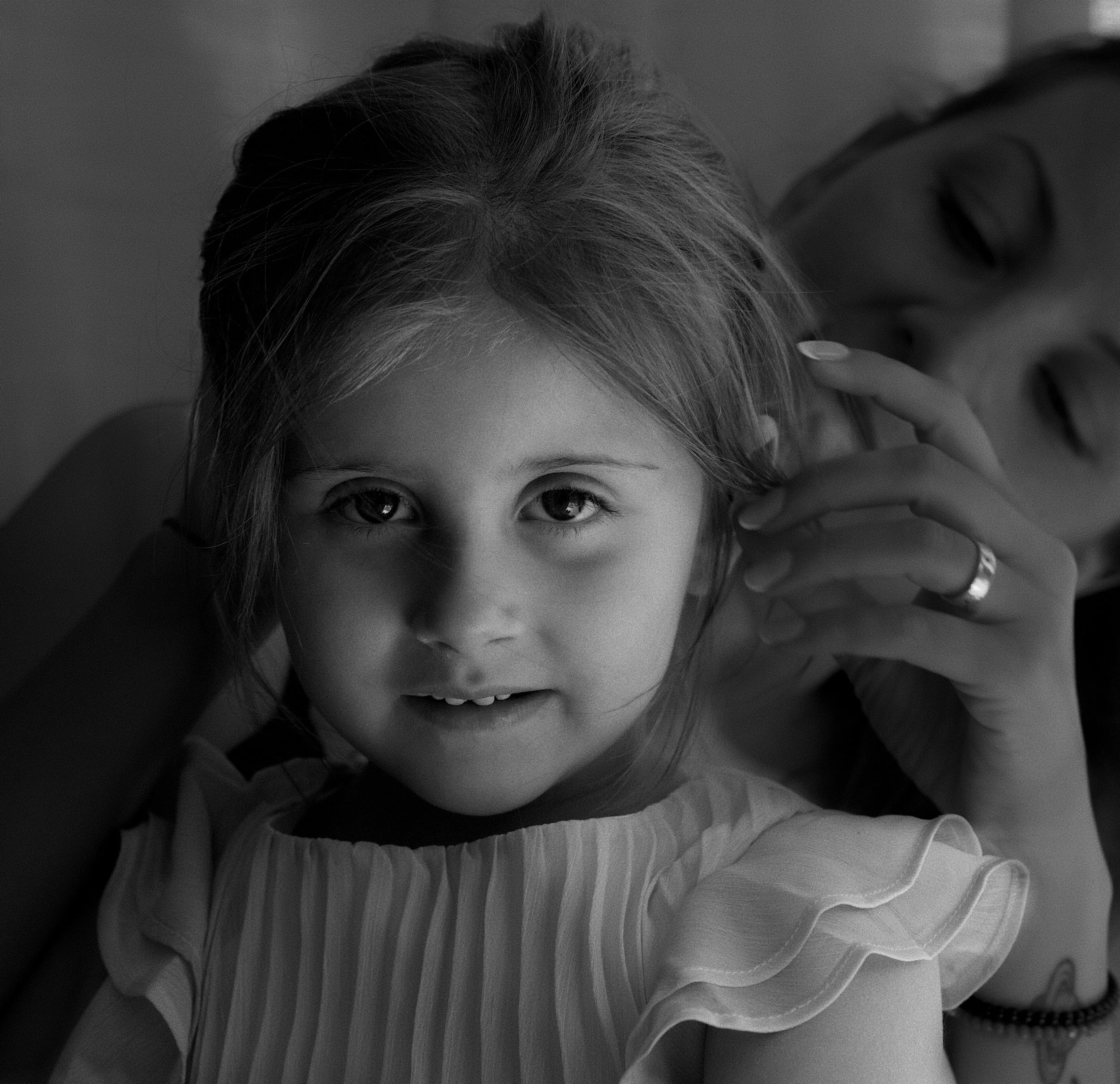 Close-up of a young girl's face with big eyes, gently smiling, with her hand touching her head, in black and white.