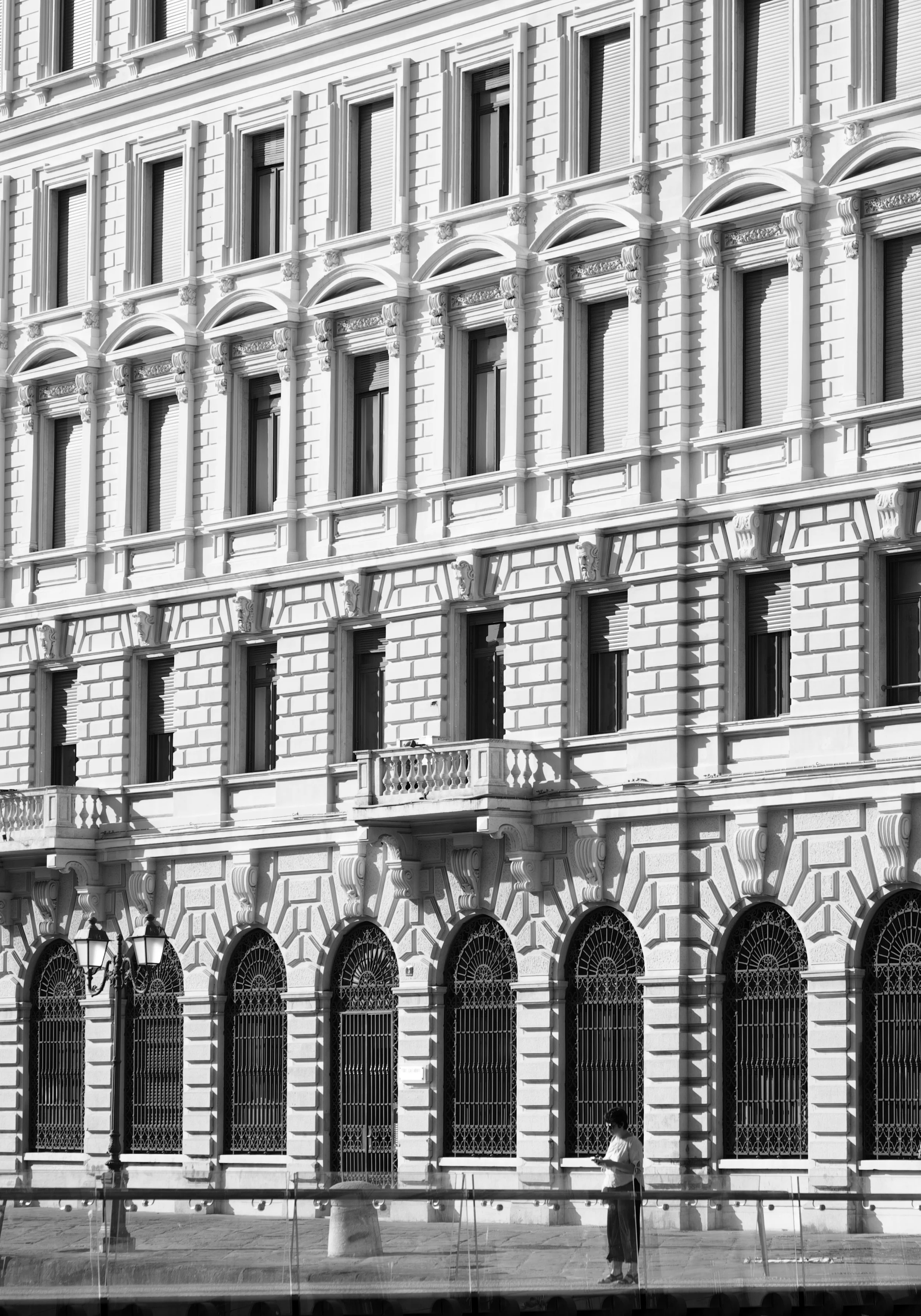 A black-and-white photo of a multi-story historic building with ornate window frames, decorative moldings, and arches on the ground floor, with a woman standing outside reading a book or notebook.