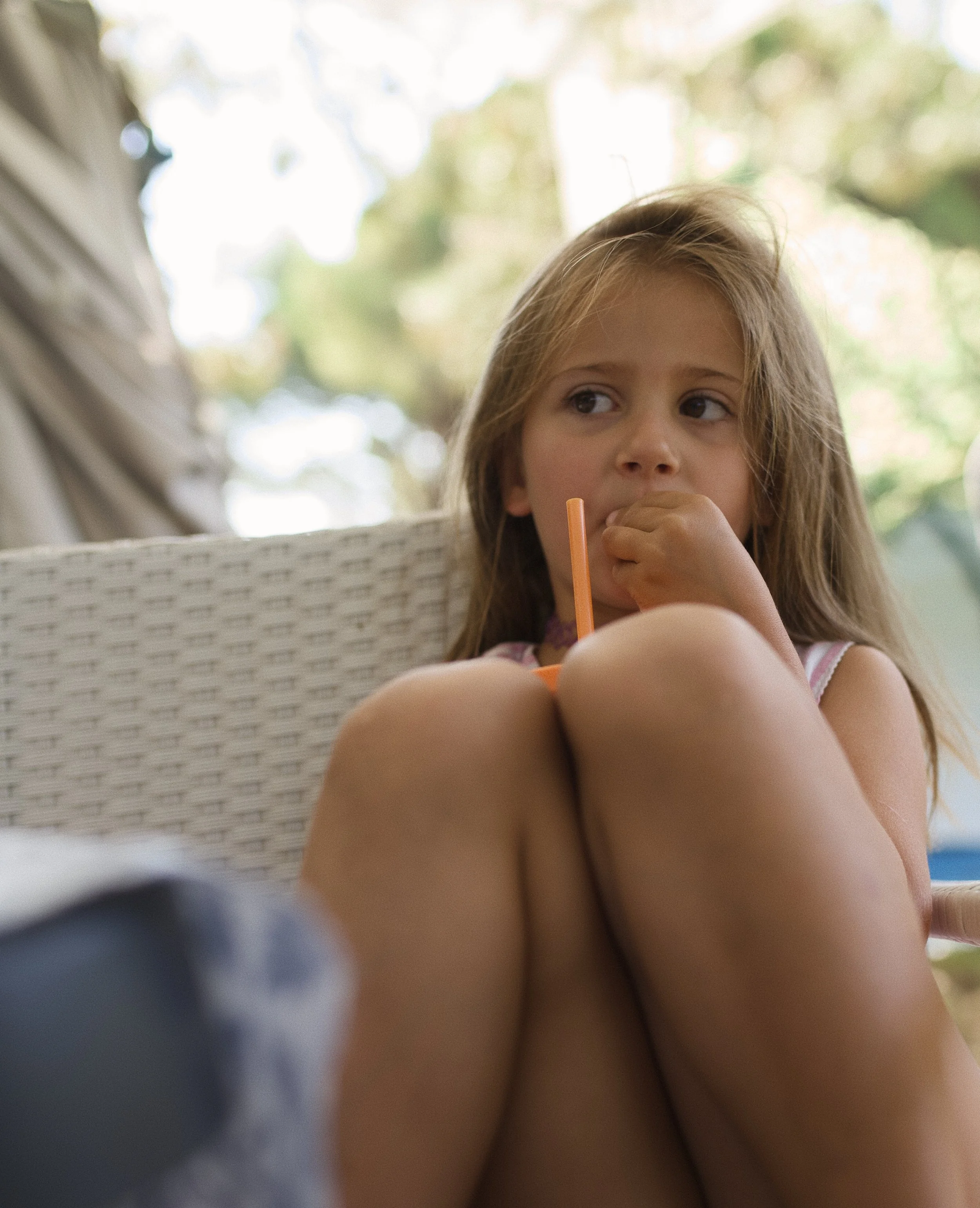 Close-up of a young girl sitting outdoors with blurred trees in the background, holding a drink with a straw.