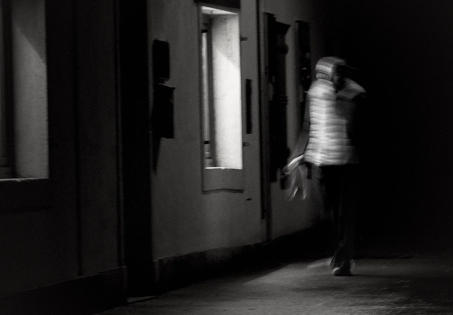 A person walking in a dark hallway, illuminated by a light coming through an open door, wearing a striped shirt and dark pants.