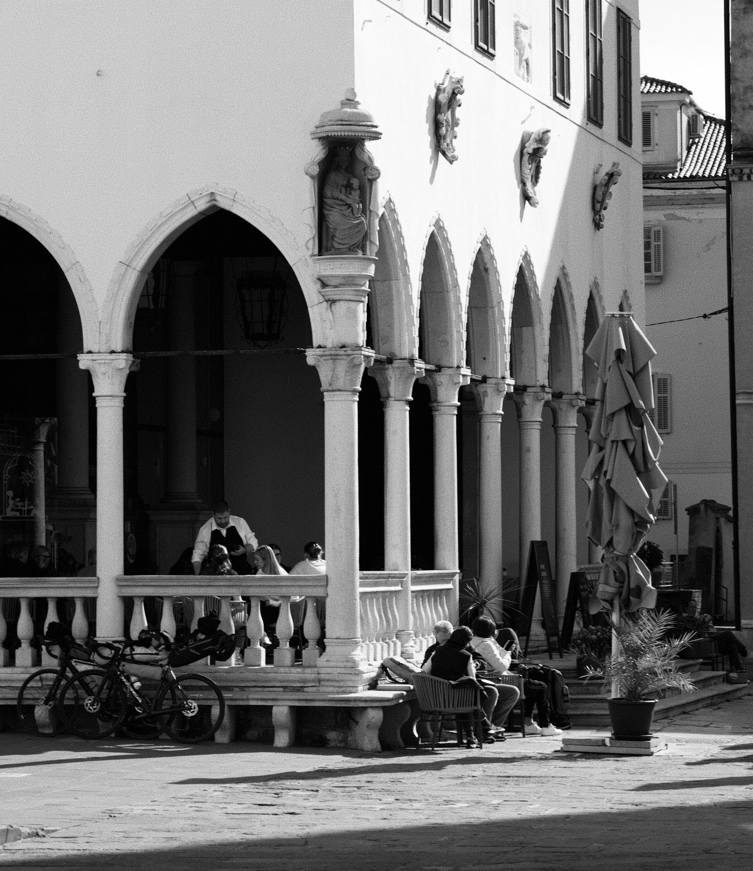A lively outdoor café with people seated at tables under a pavilion with arched openings and decorative statues on the facade. Bicycle stands are in the foreground, and there are large umbrellas on the right. The building in the background has multip