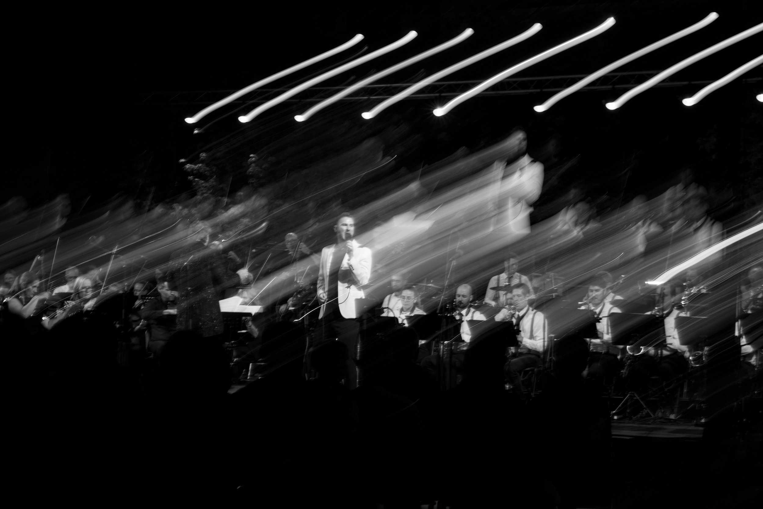 Black and white photo of a live musical performance featuring a male singer holding a microphone and band members playing instruments on stage with abstract light streaks in the background.
