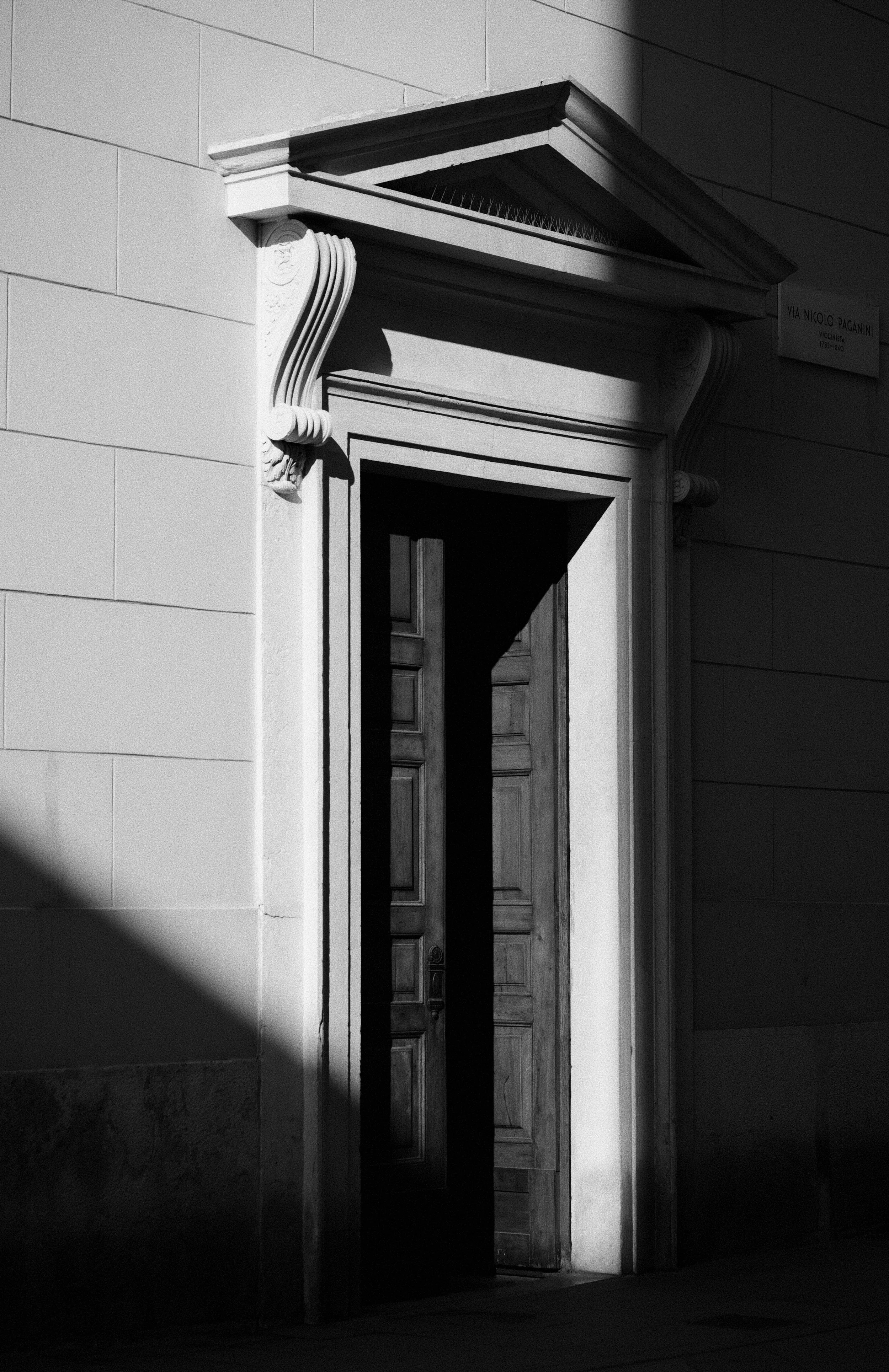 Black and white photograph of a doorway with ornate architectural details on the columns and a sign that reads 'VIA NICOLO PAGANINI VOLORECICA 1733-1840' on the wall adjacent to the door.