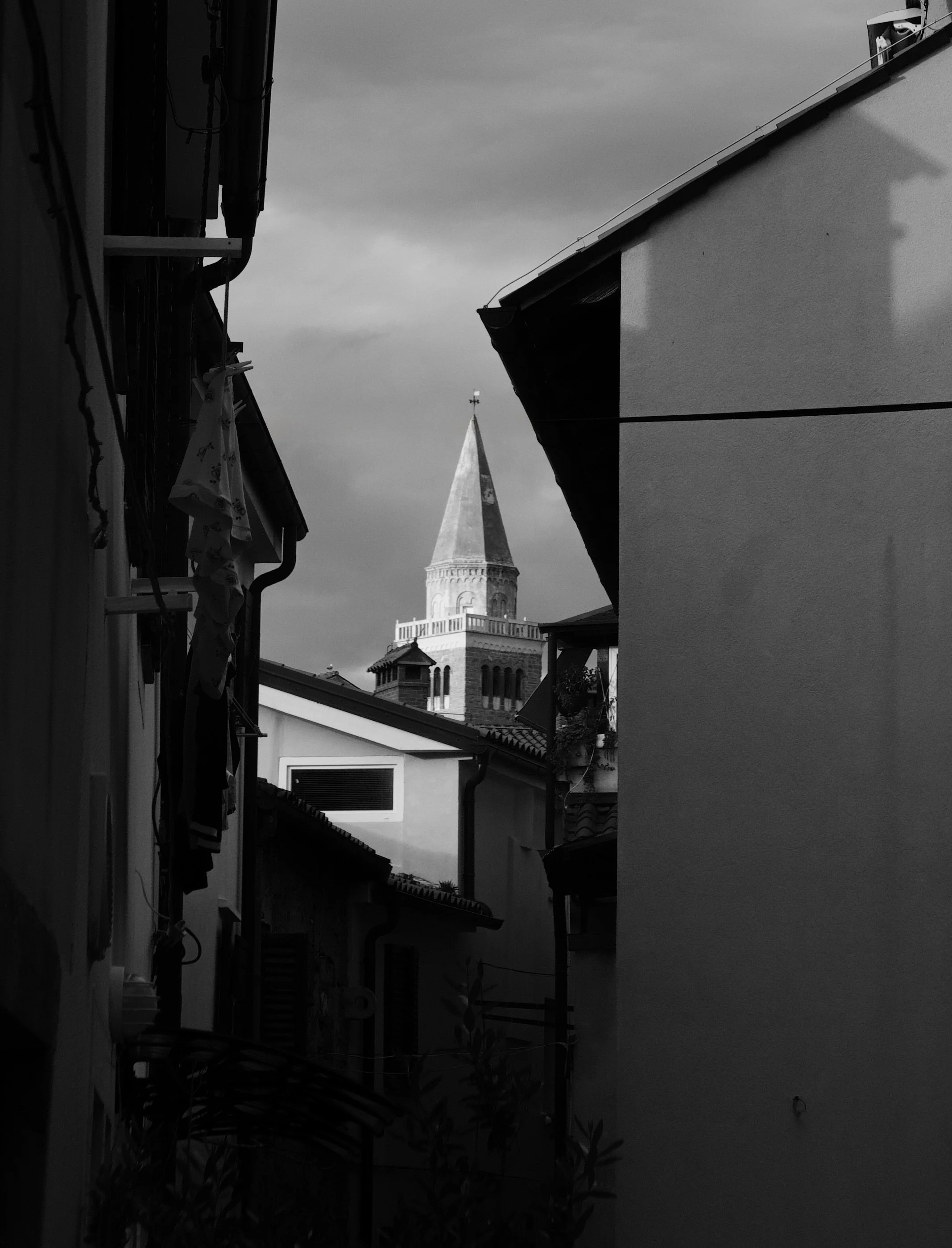 View of a church steeple with a weather vane, framed by the roofs of nearby buildings, under a cloudy sky.