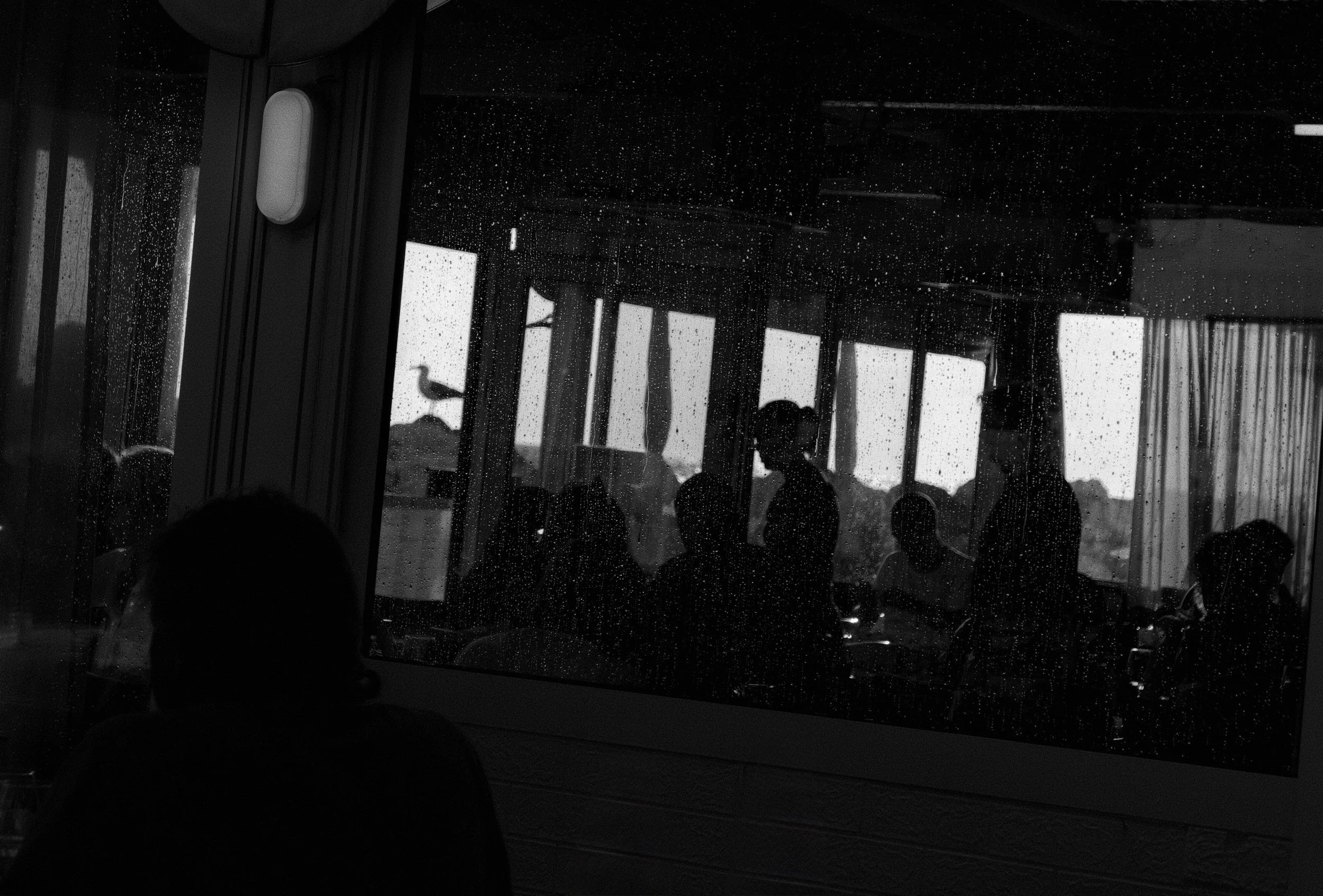 The image depicts an indoor scene viewed through a rain-covered window, with silhouettes of people gathered and some standing near the windows.