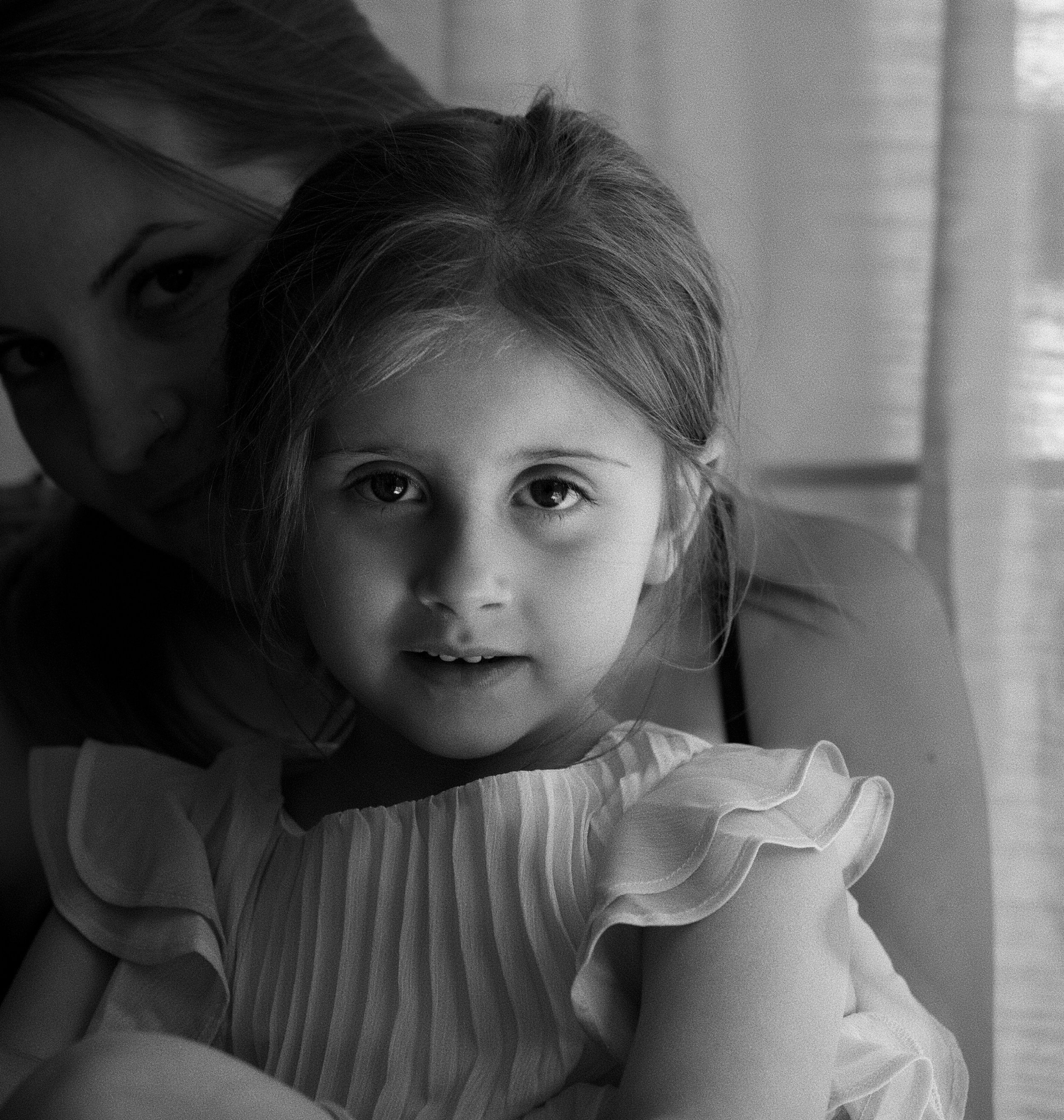 Black and white photo of a woman and a young girl close together, both smiling and looking at the camera. The woman appears to be lying on her side, and the girl is positioned in front of her. The background shows window blinds.