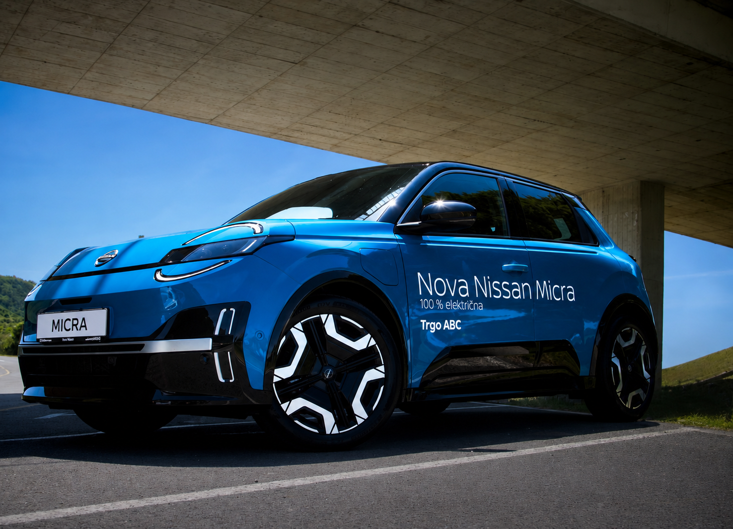 Blue Nissan Micra electric vehicle parked under a concrete overpass with green hills in the background.