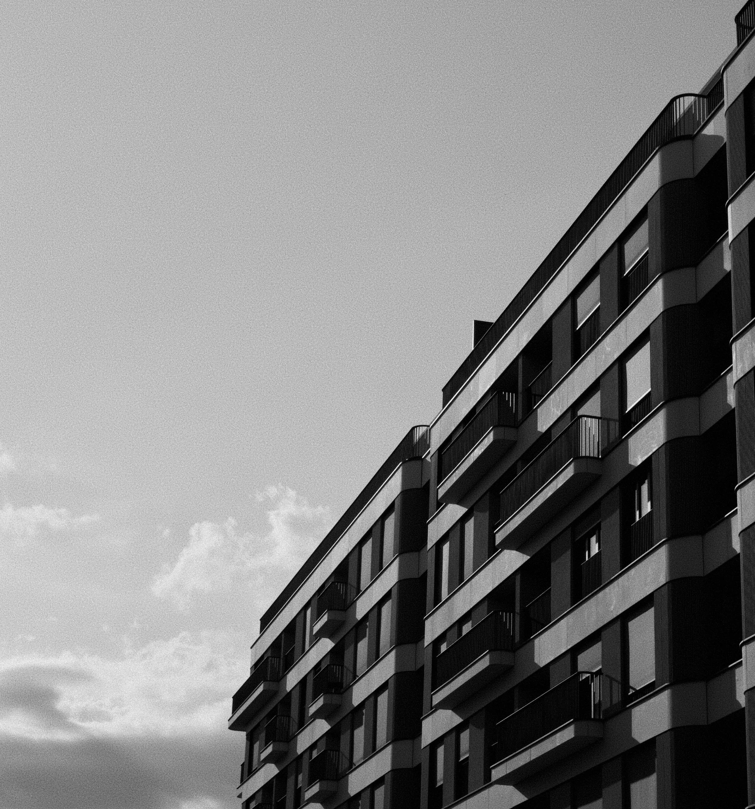 A black and white photo of a modern multi-story building with curved balconies and large glass windows, set against a partly cloudy sky.