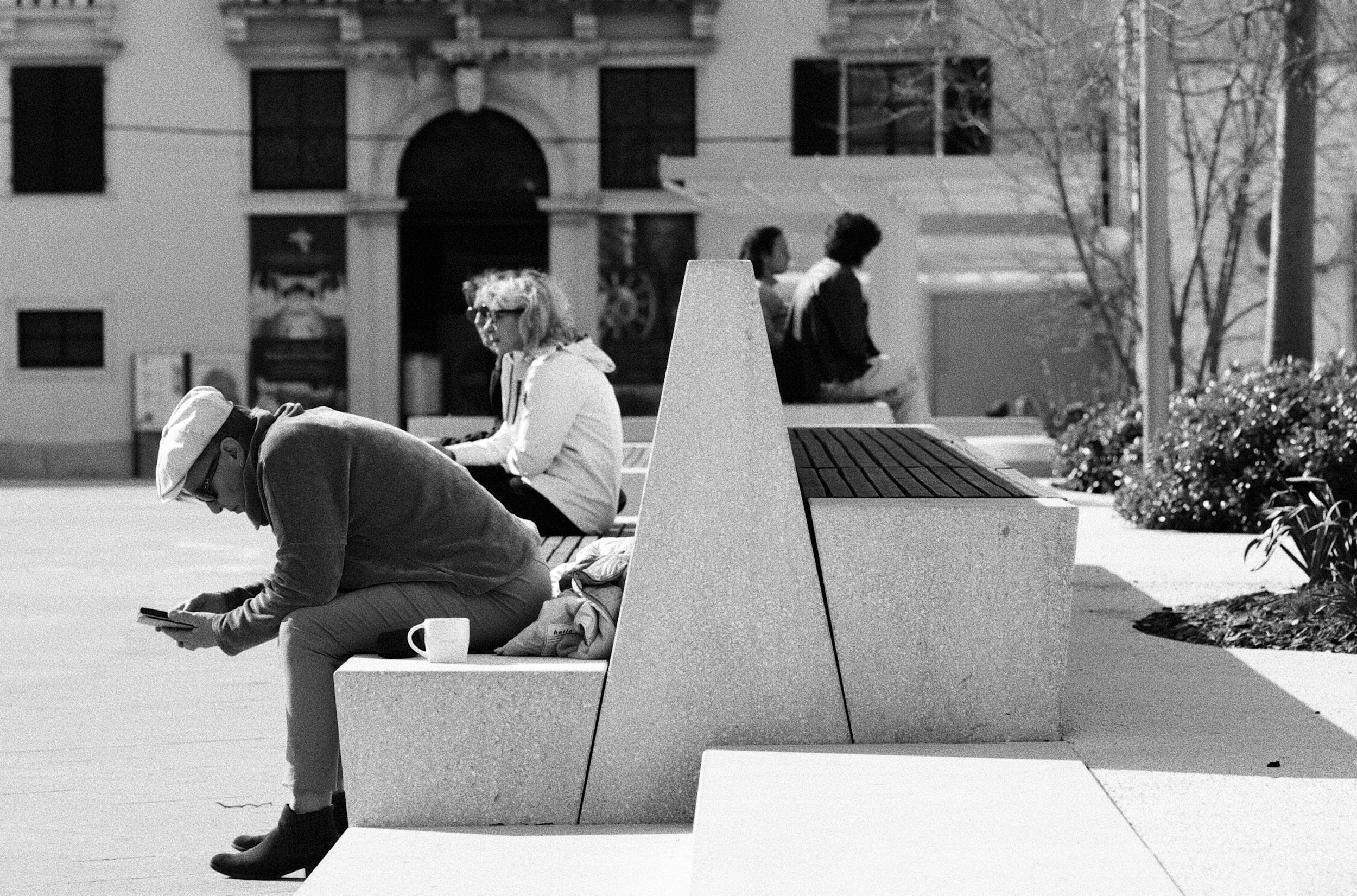 Three people sitting on benches outdoors, two women and one man, with buildings and trees in the background, black and white photo.
