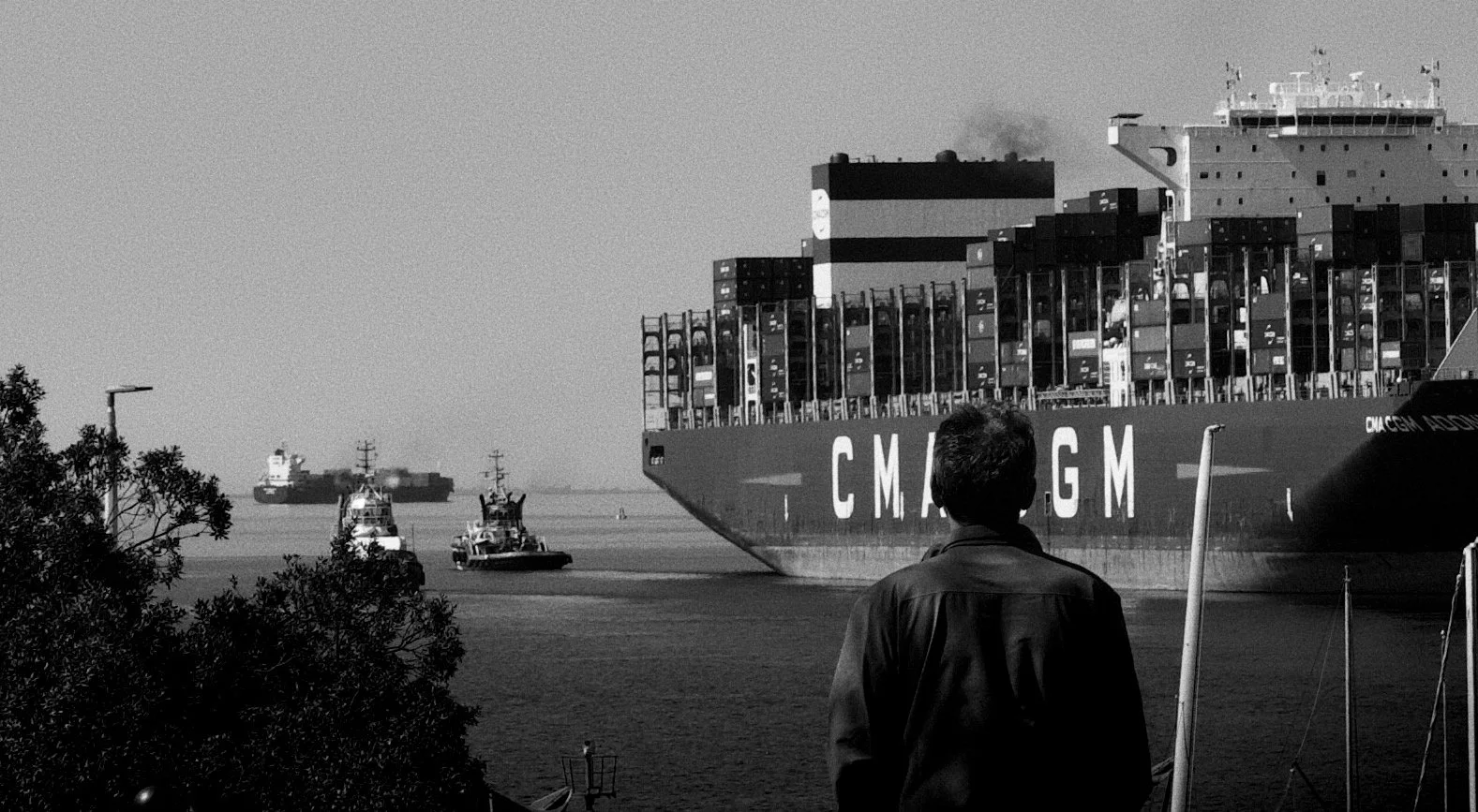 A man standing on the shore, observing a large container ship with the letters 'CMA CGM' on its side, sailing in the water, with smaller boats nearby.