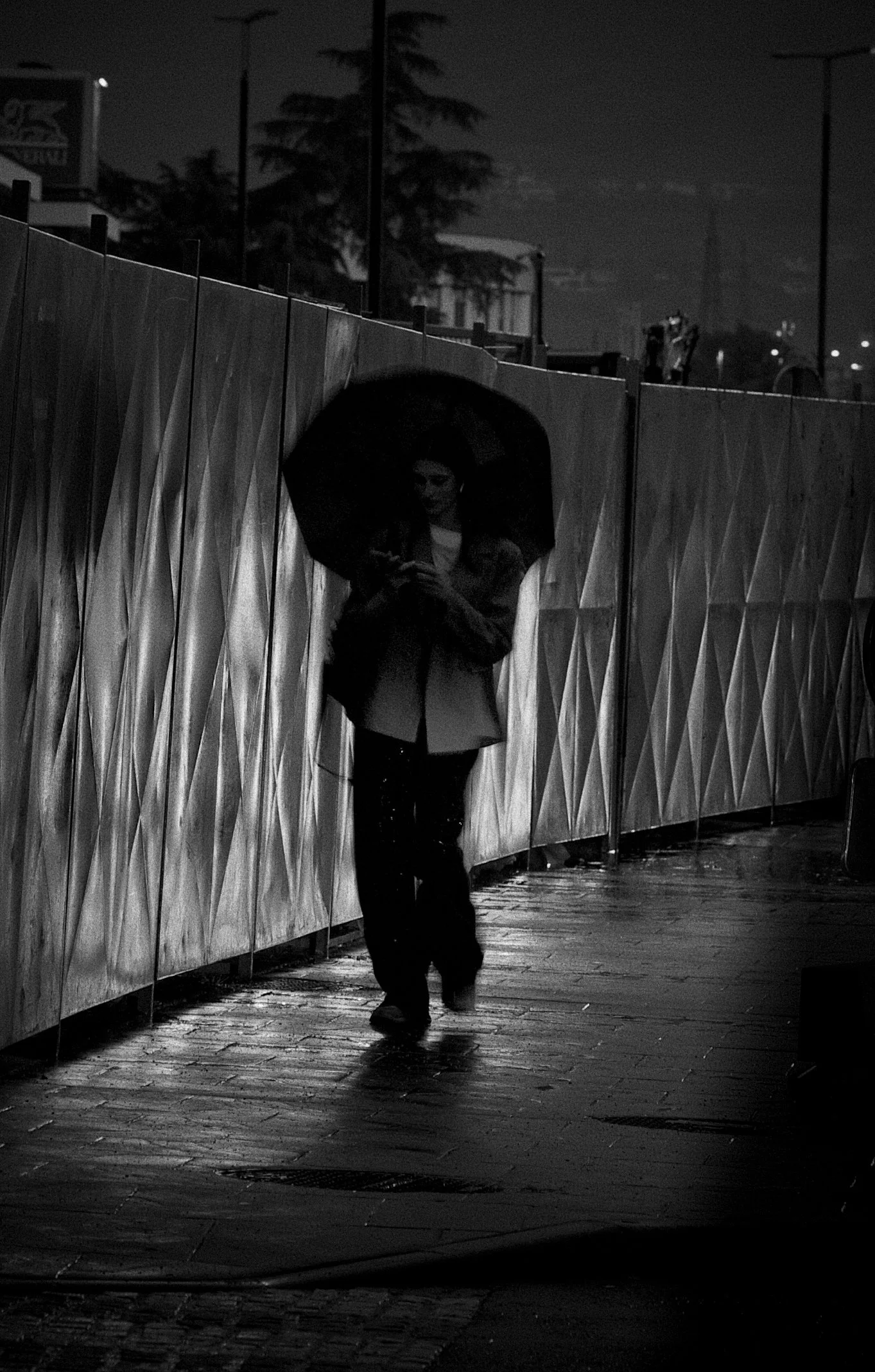 A woman walking with an umbrella at night behind a metal fence, wet pavement, and city lights in the background.