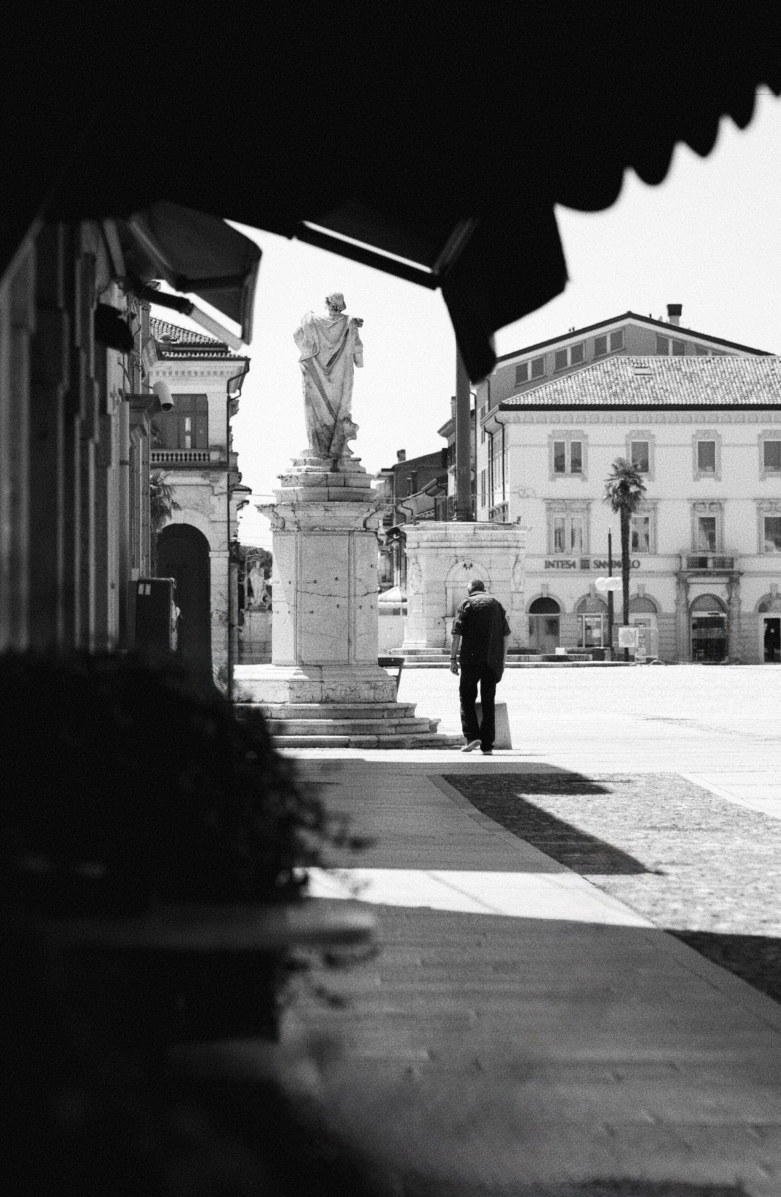 A black and white street scene viewed through an opening, showing a man walking near a statue of a robed figure on a column in the square. The background features classic European-style buildings.