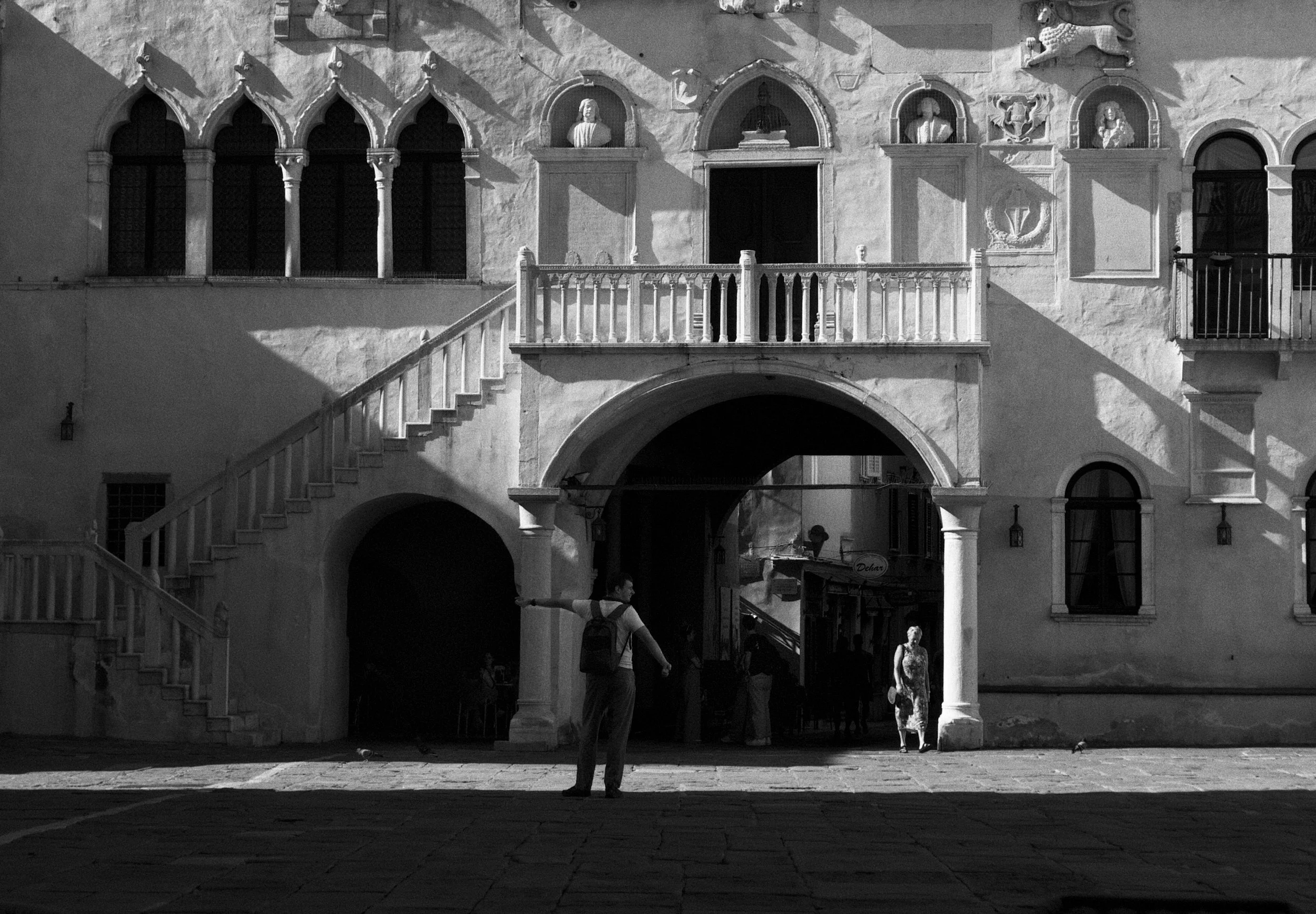 A black-and-white photo of a building with gothic and classical architectural features, including arched windows, columns, and statues. A staircase leads up to a balcony, and a man with a backpack is standing in the foreground, holding something in h