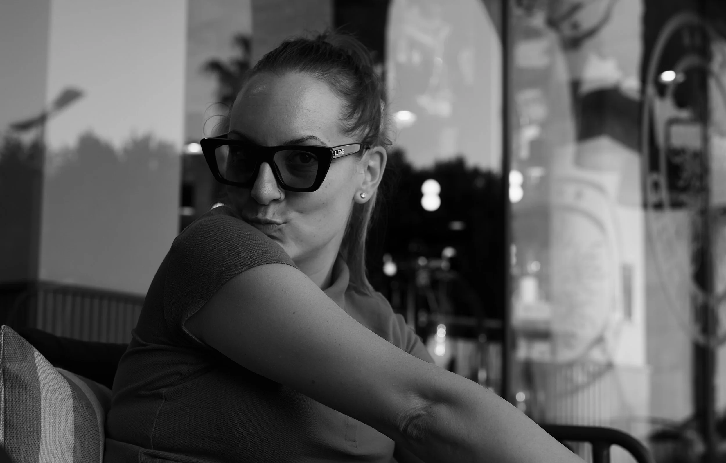 Black and white photo of a young woman with glasses and a nose piercing, sitting indoors with a contemplative expression, reflected in a window with a cityscape background.