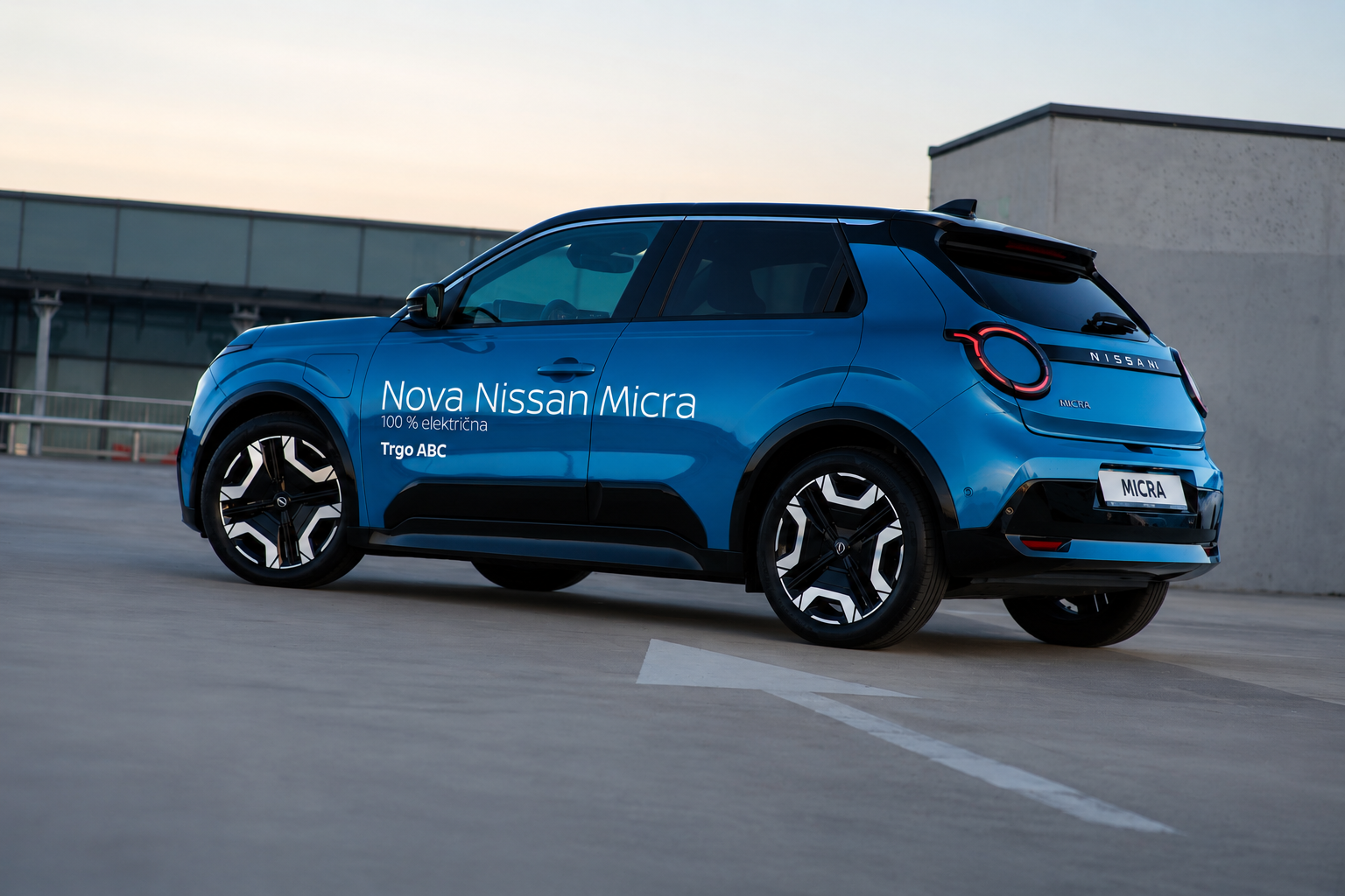 Blue Nissan Micra electric car parked on a rooftop with promotional text on its side reading "Nova Nissan Micra, 100% električna, Trgo ABC."