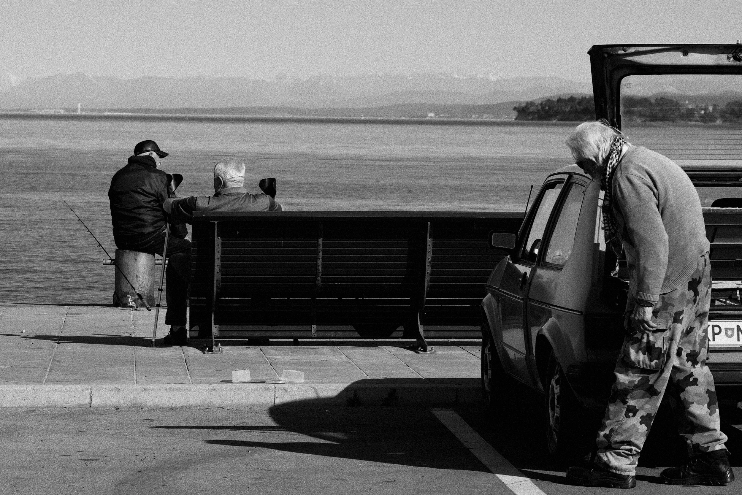 Two elderly men fishing off a pier while a woman stands near a parked vehicle on the street. The scene is by the water with distant land and mountains in the background.