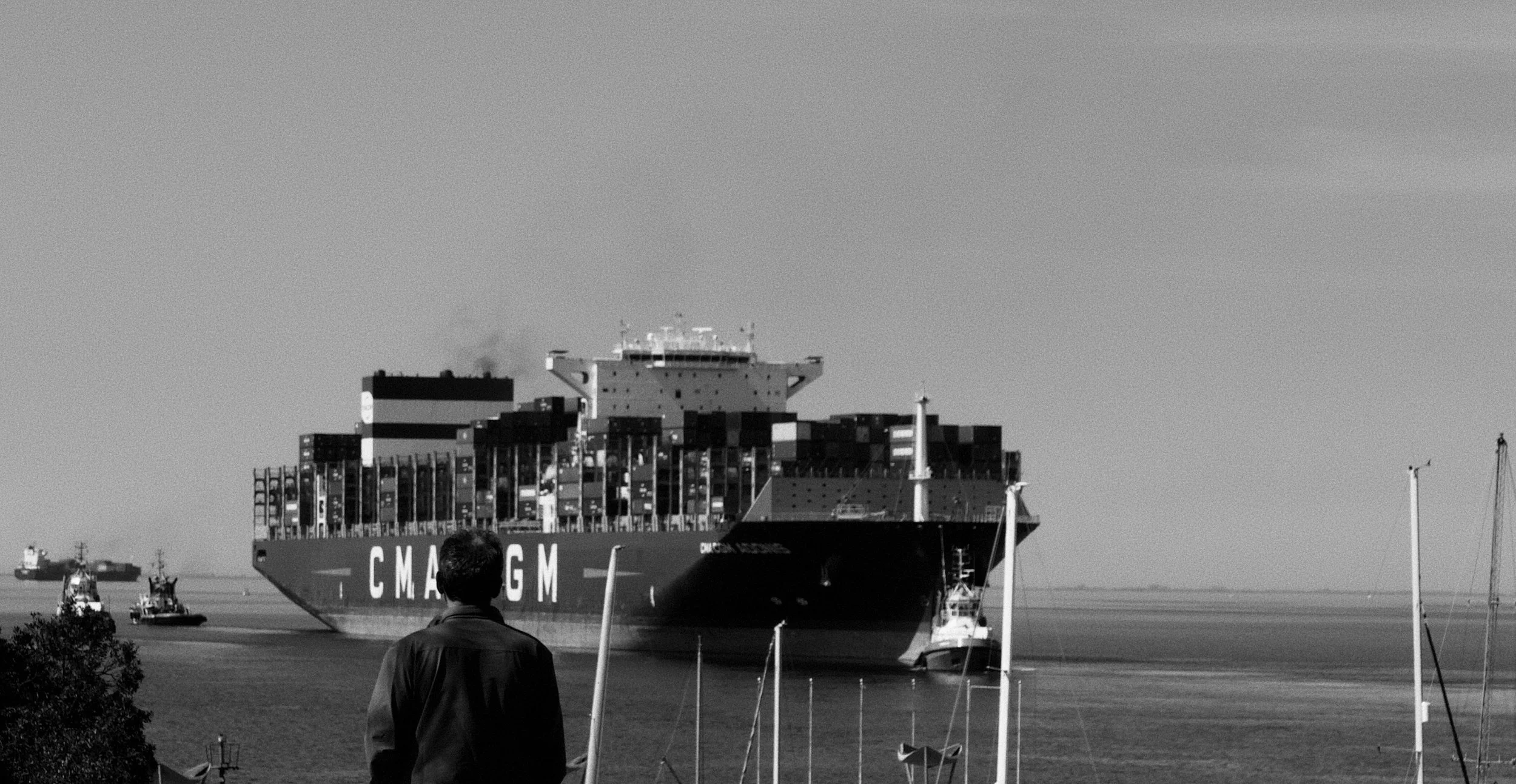 A large container ship labeled 'CMA CGM' sailing in a harbor with smaller boats and a person in the foreground, black and white photo.