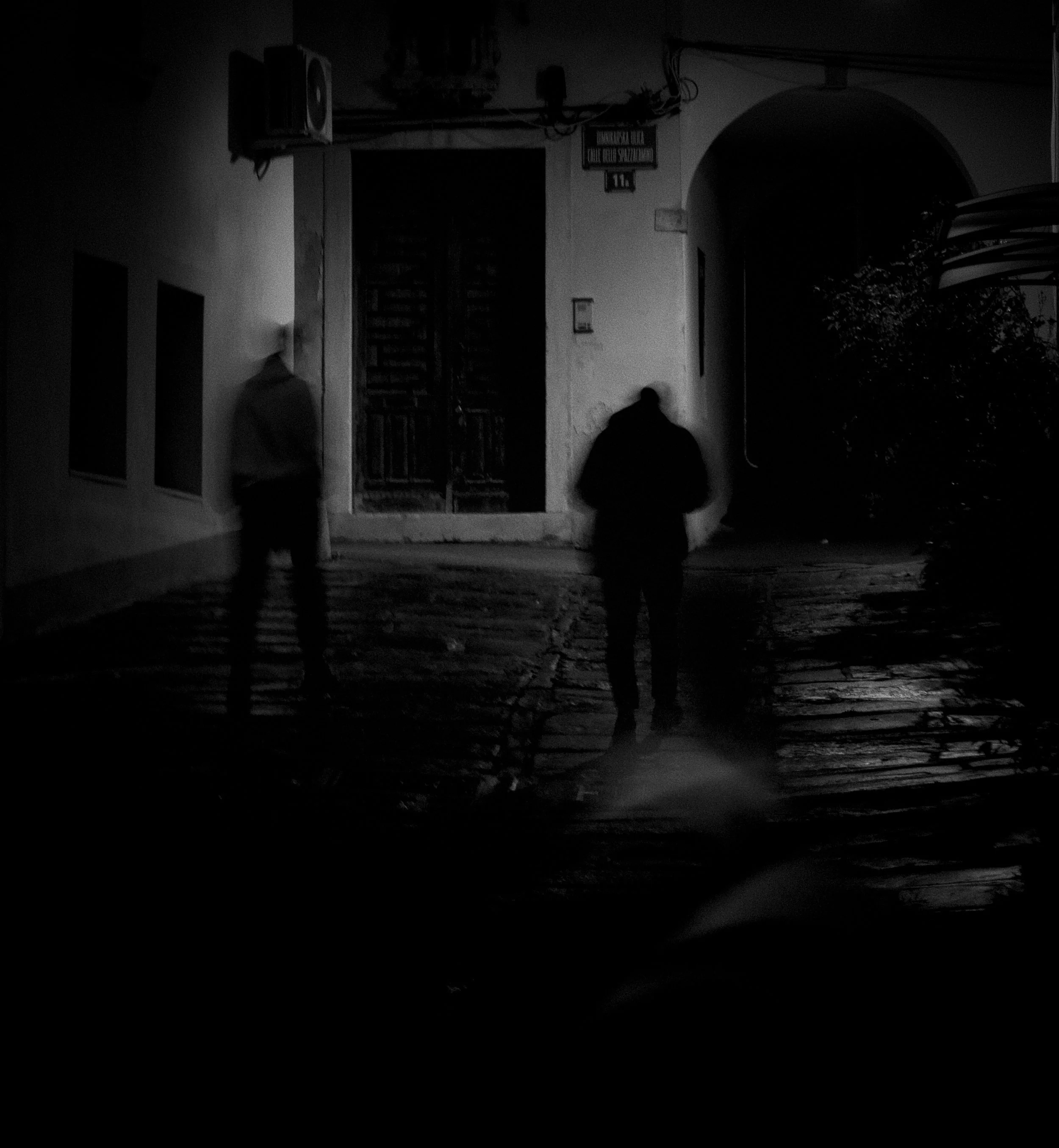 Dark photo of a building entrance with three silhouetted figures walking in front. The building has a sign with unreadable text and a staircase visible behind a gate.