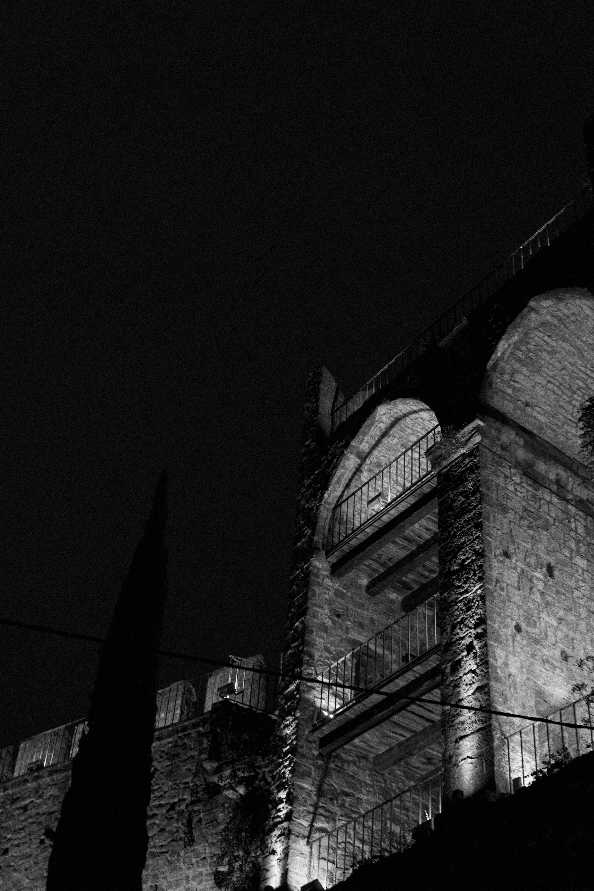 Nighttime view of an old brick building with stairs and a railing, illuminated by spotlights, against a dark sky with a tall, shadowed tree nearby.