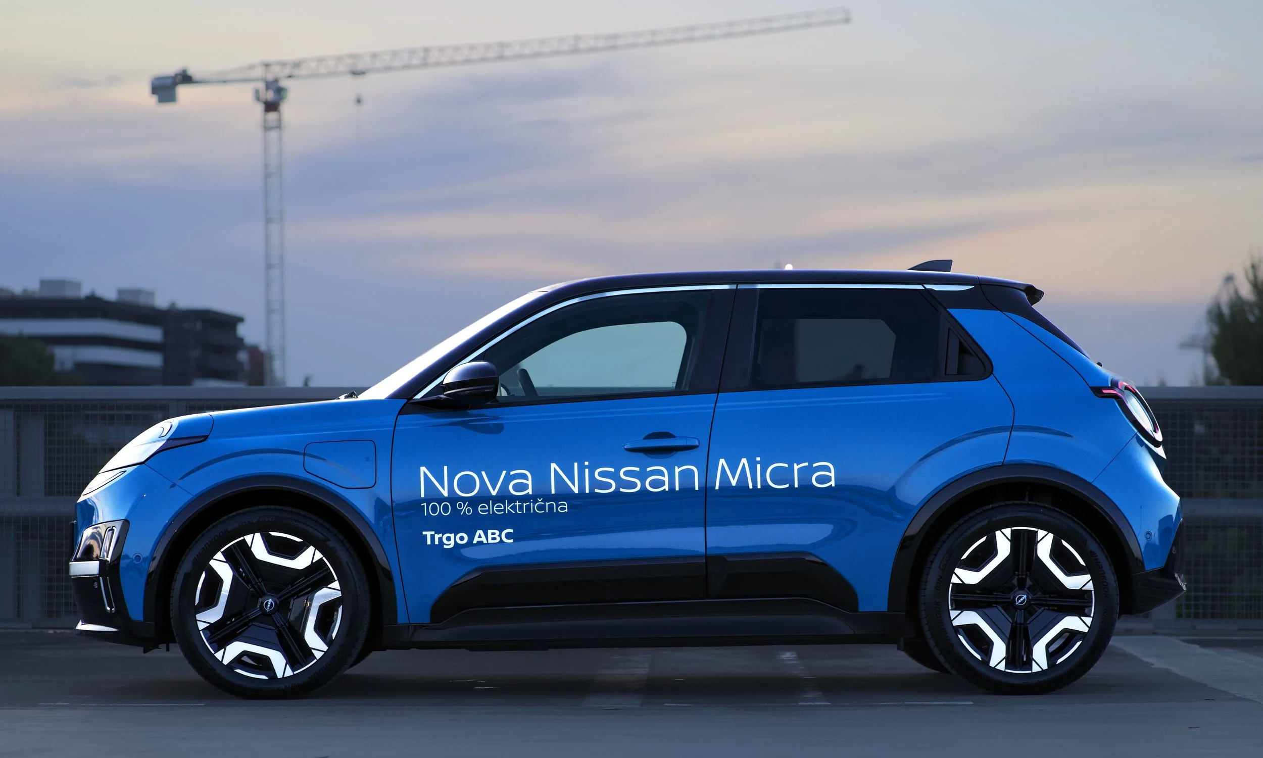 A blue Nissan Micra electric car parked outdoors during sunset with construction crane and buildings in the background.
