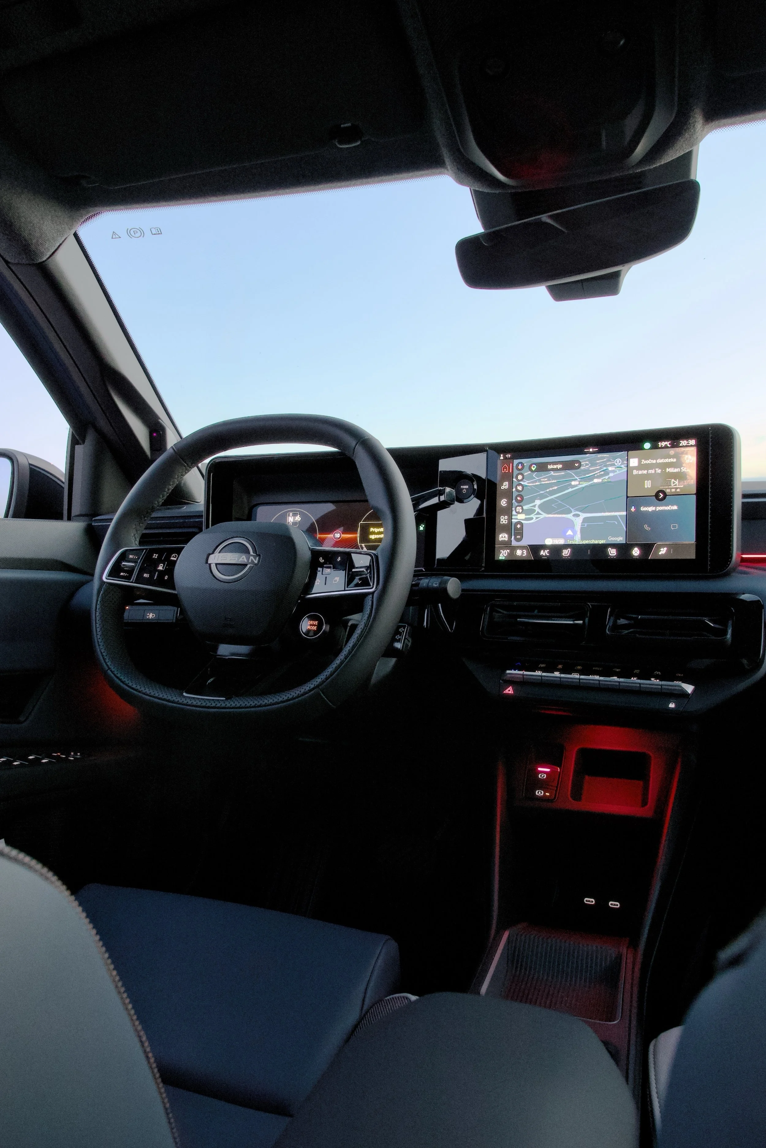 The interior of a Nissan vehicle showing the steering wheel, digital dashboard, and a large touchscreen display with a navigation map. The sky is visible through the windshield.