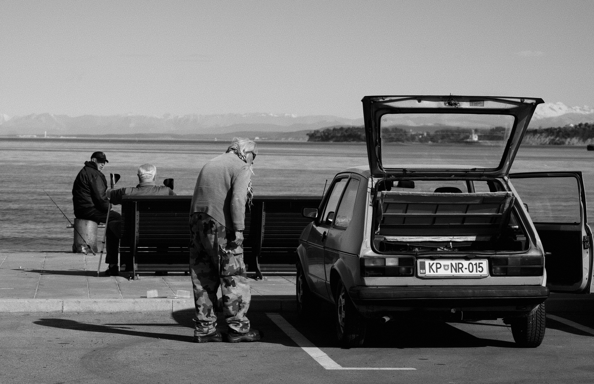 Three people near a parked car by the water, two sitting and one standing, with a view of distant mountains and shoreline in the background.