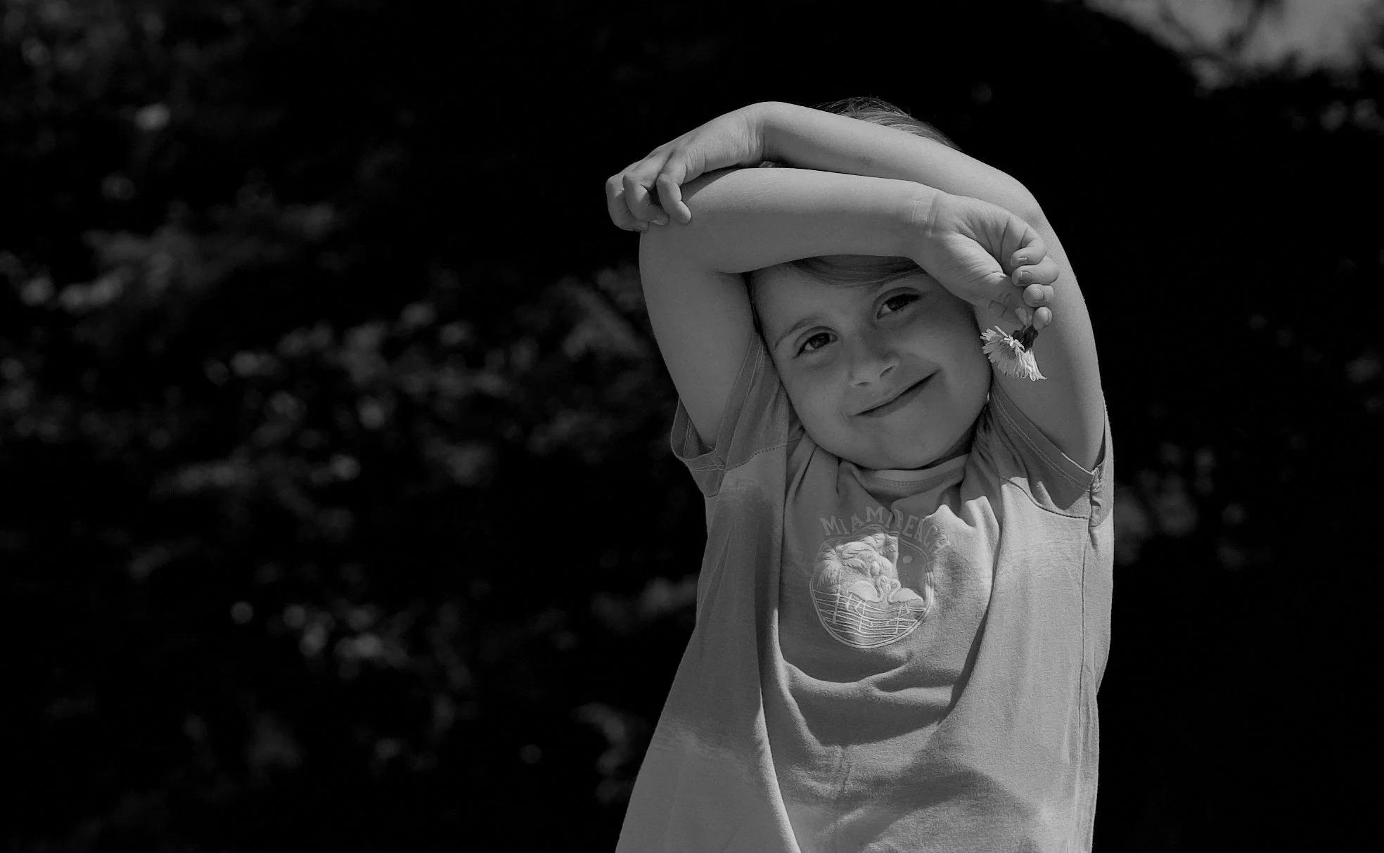 A young girl smiling with her arms raised overhead, holding a daisy flower, outdoors with a blurred dark background.