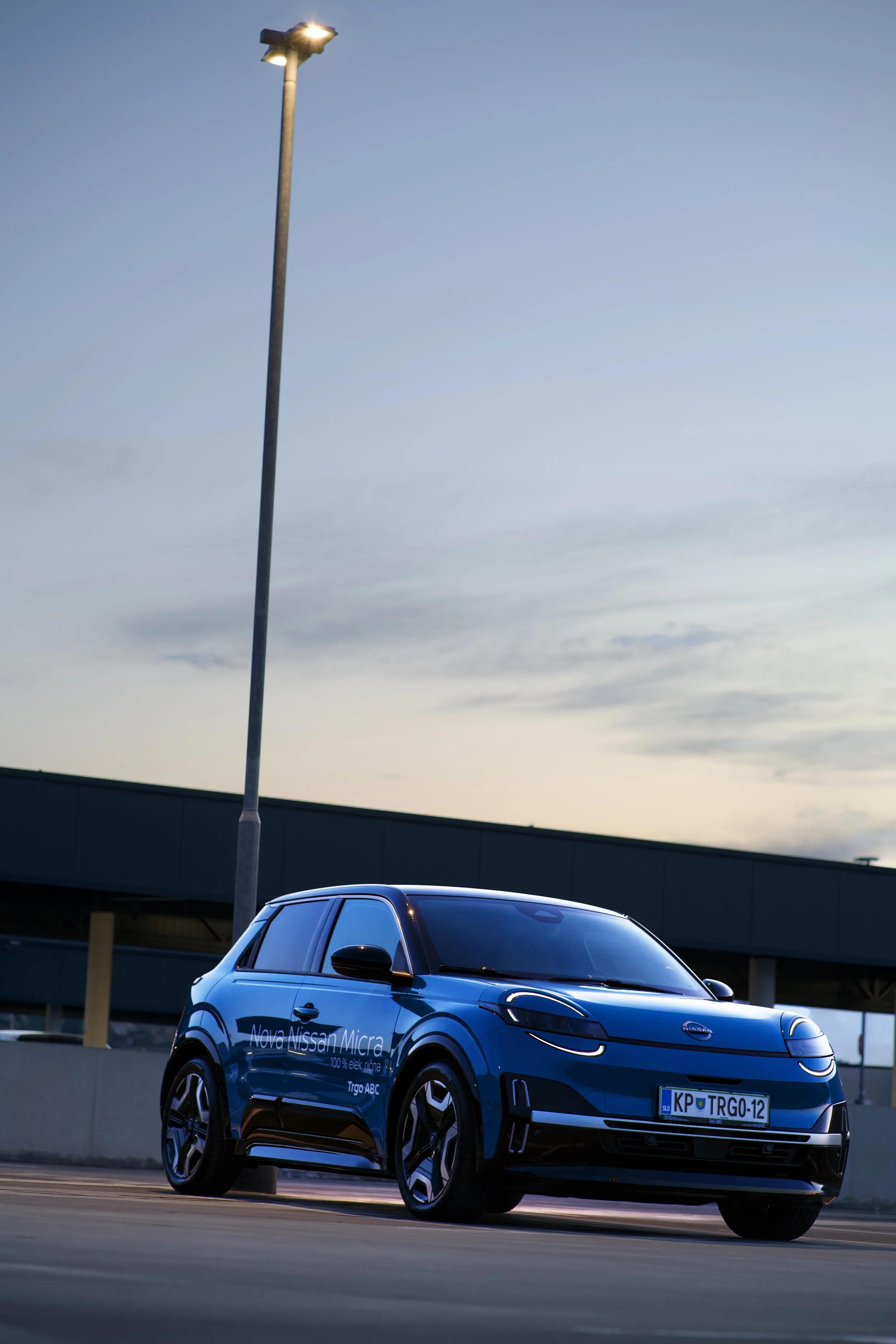 Blue electric Nissan Micra car parked in a lot, with a tall street light and a building in the background during dusk.