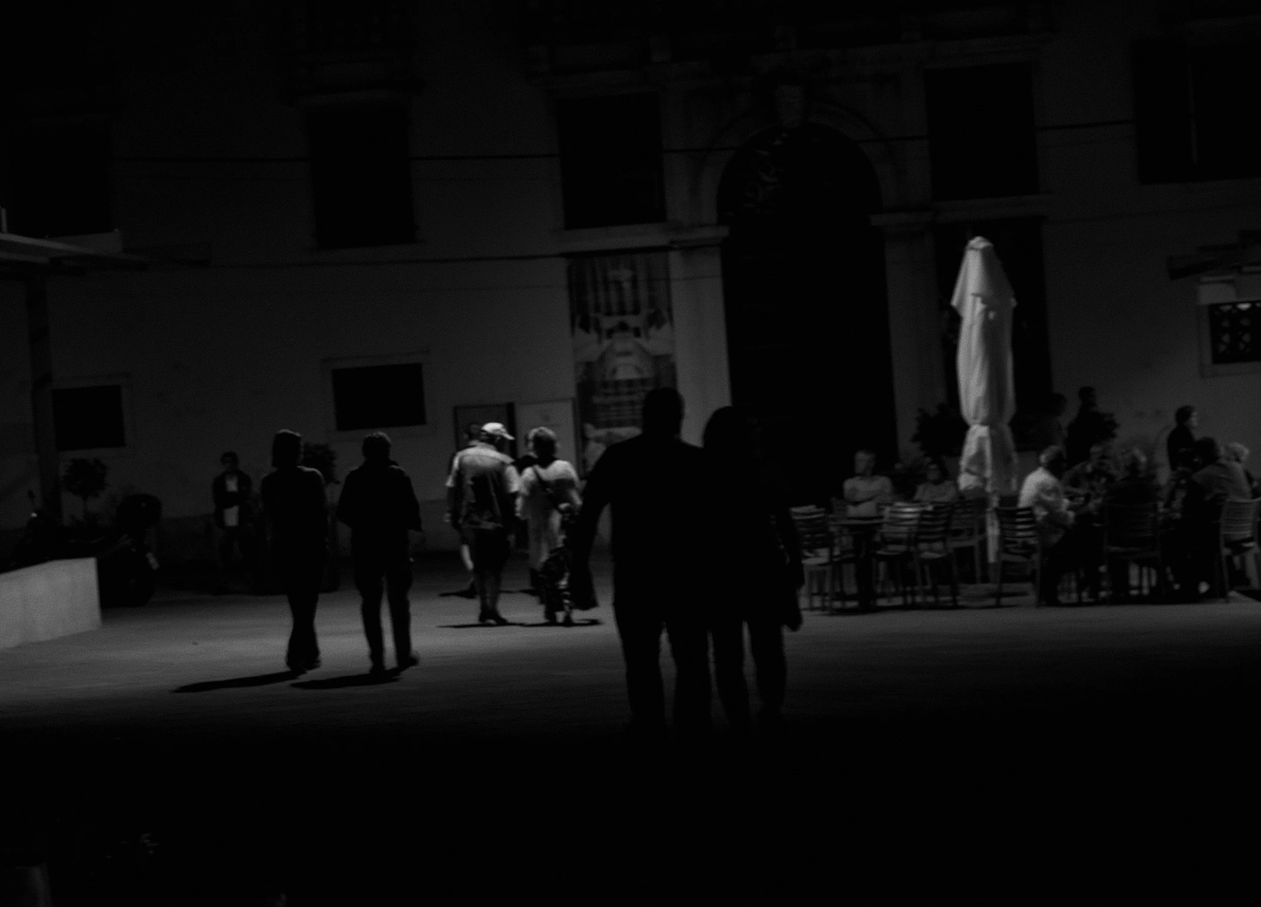 People gathered outdoors at night with a building in the background, some tables and an umbrella visible, illuminated by a light source.
