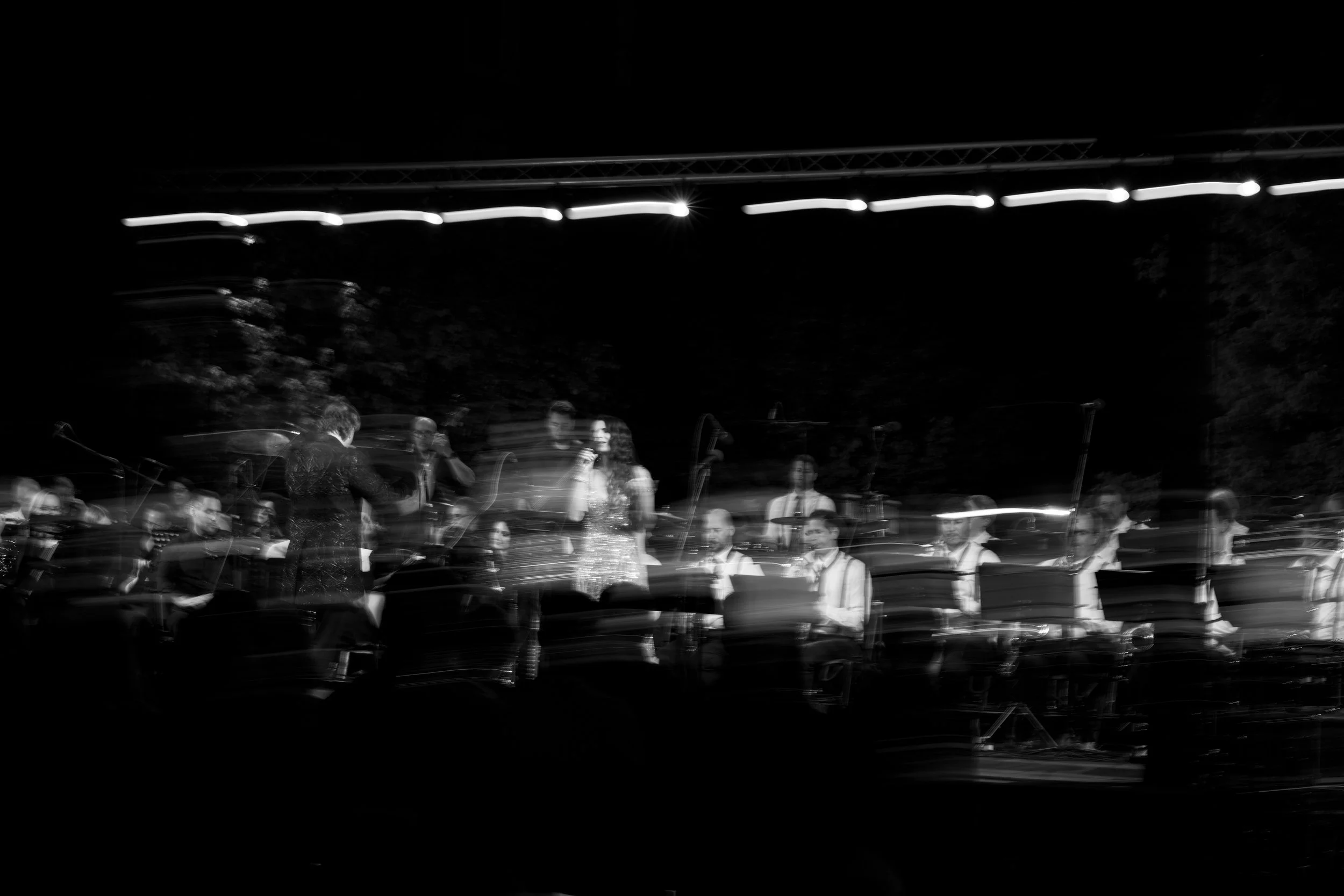 A black-and-white photo of a night scene with a group of musicians performing outdoors, with some wearing suspenders and ties. String lights are hung above them, and there are trees in the background.