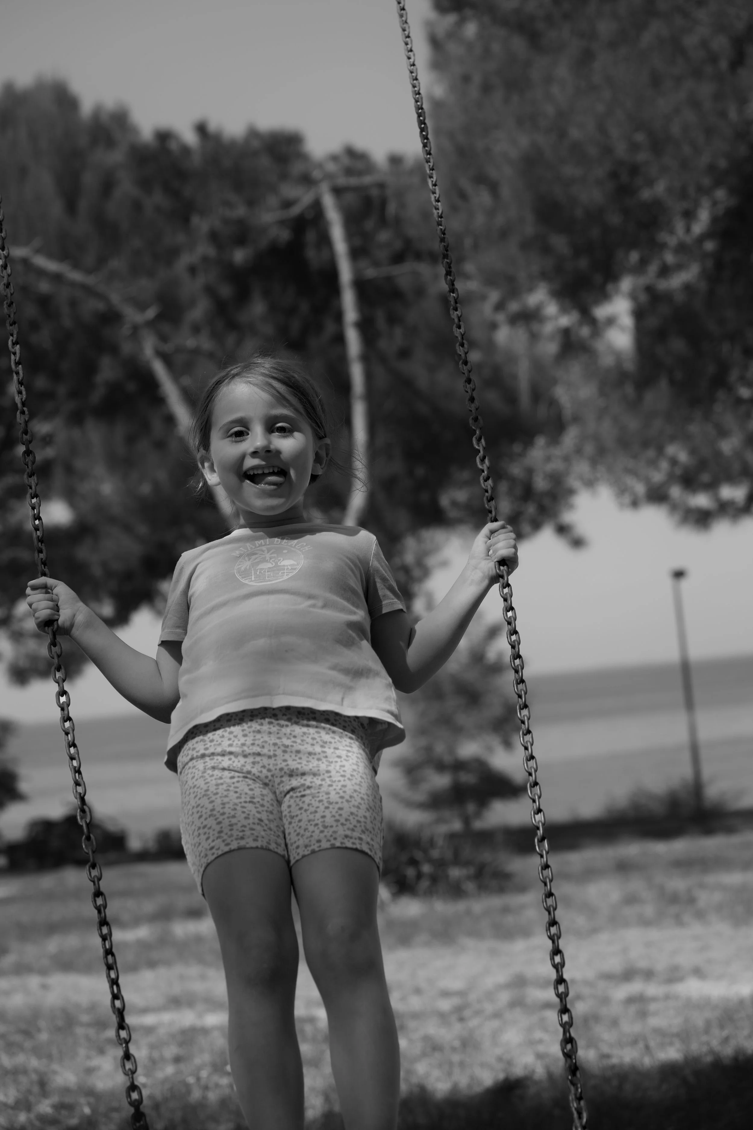 A young girl with a big smile and tongue out, swinging on a playground set with chains, in a park with trees and grass in the background, in black and white.