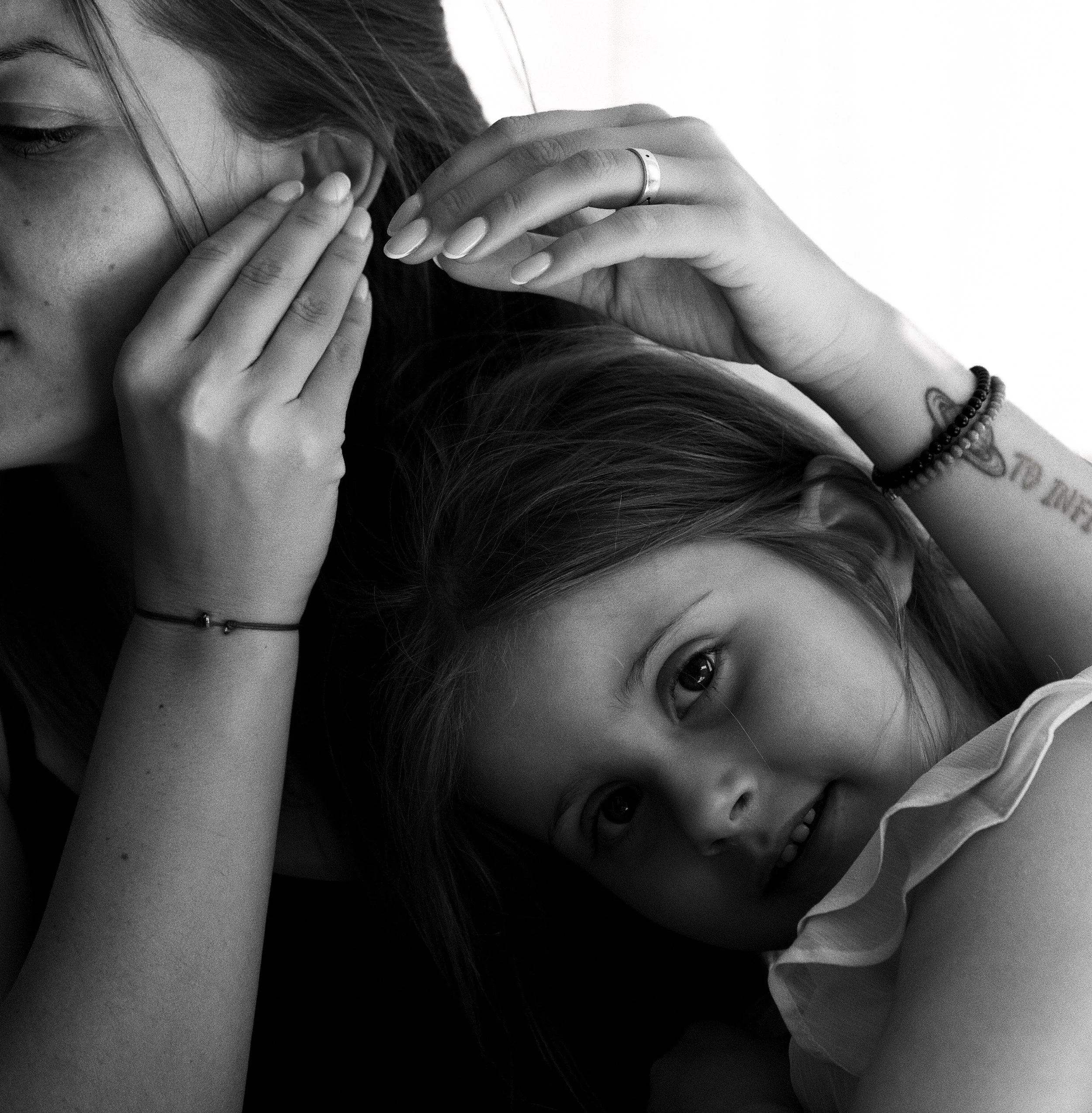 Black and white close-up photo of a woman with long hair, partially covering her face, eyes gazing downward, hands resting on her face, wearing a bracelet on her wrist, and a ring on her finger.