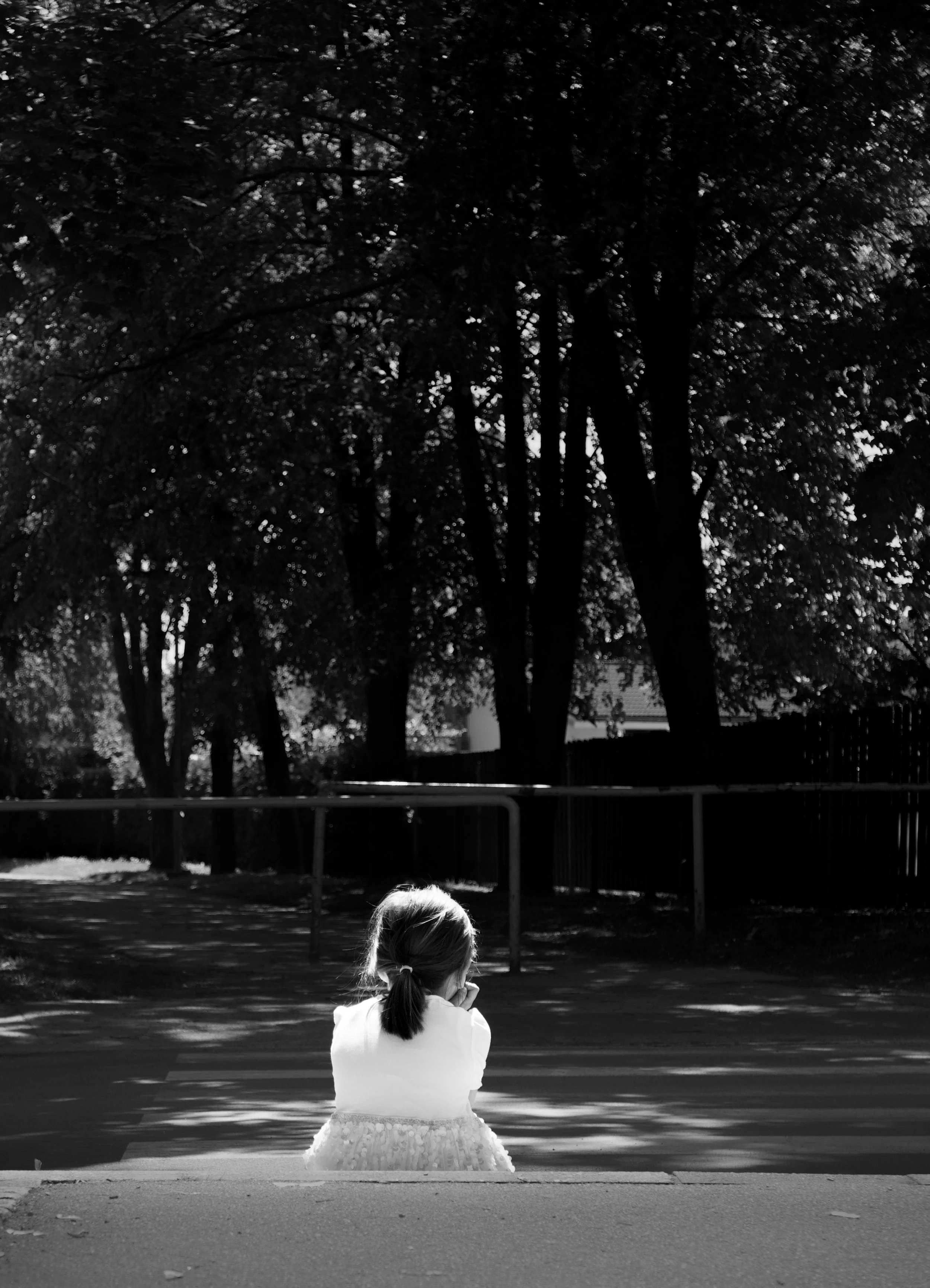 A young girl with pigtails wearing a dress sitting on a curb, facing away, during daytime, with a background of tall trees and a fence, in black and white.