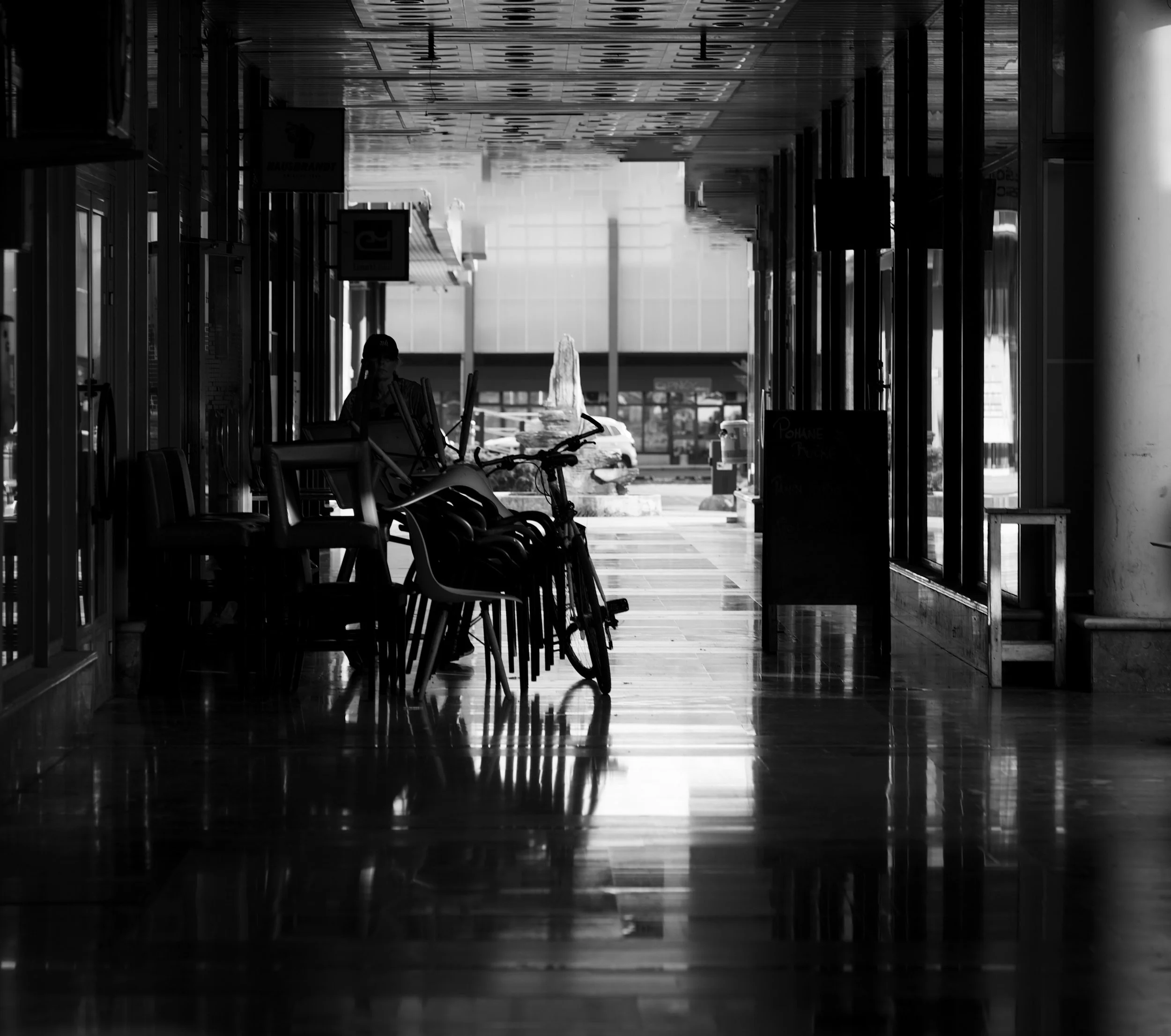 A black and white photograph of an indoor area, possibly an airport or train station, with chairs, bikes, and people in the distance near large windows and glass doors.