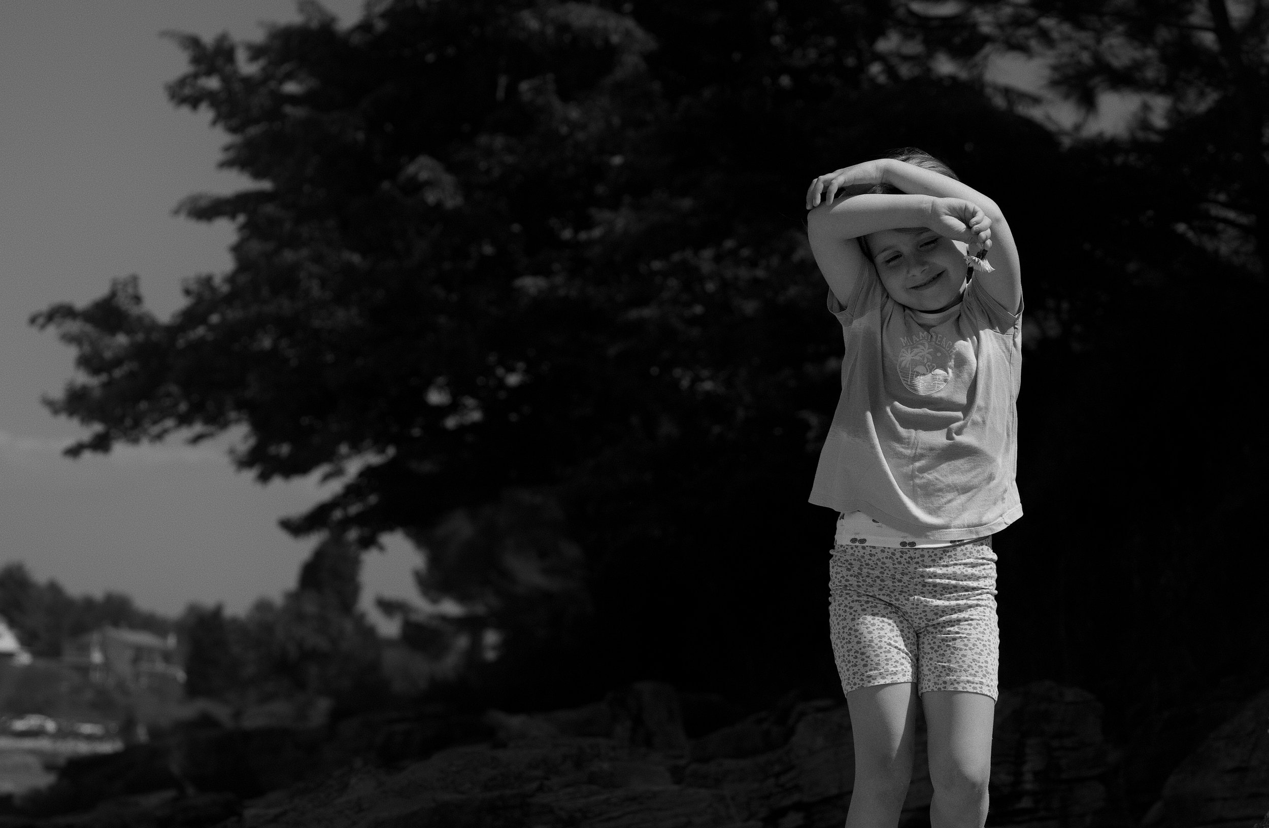 A young girl stretching outdoors near trees with a smile on her face, wearing a t-shirt and patterned leggings.