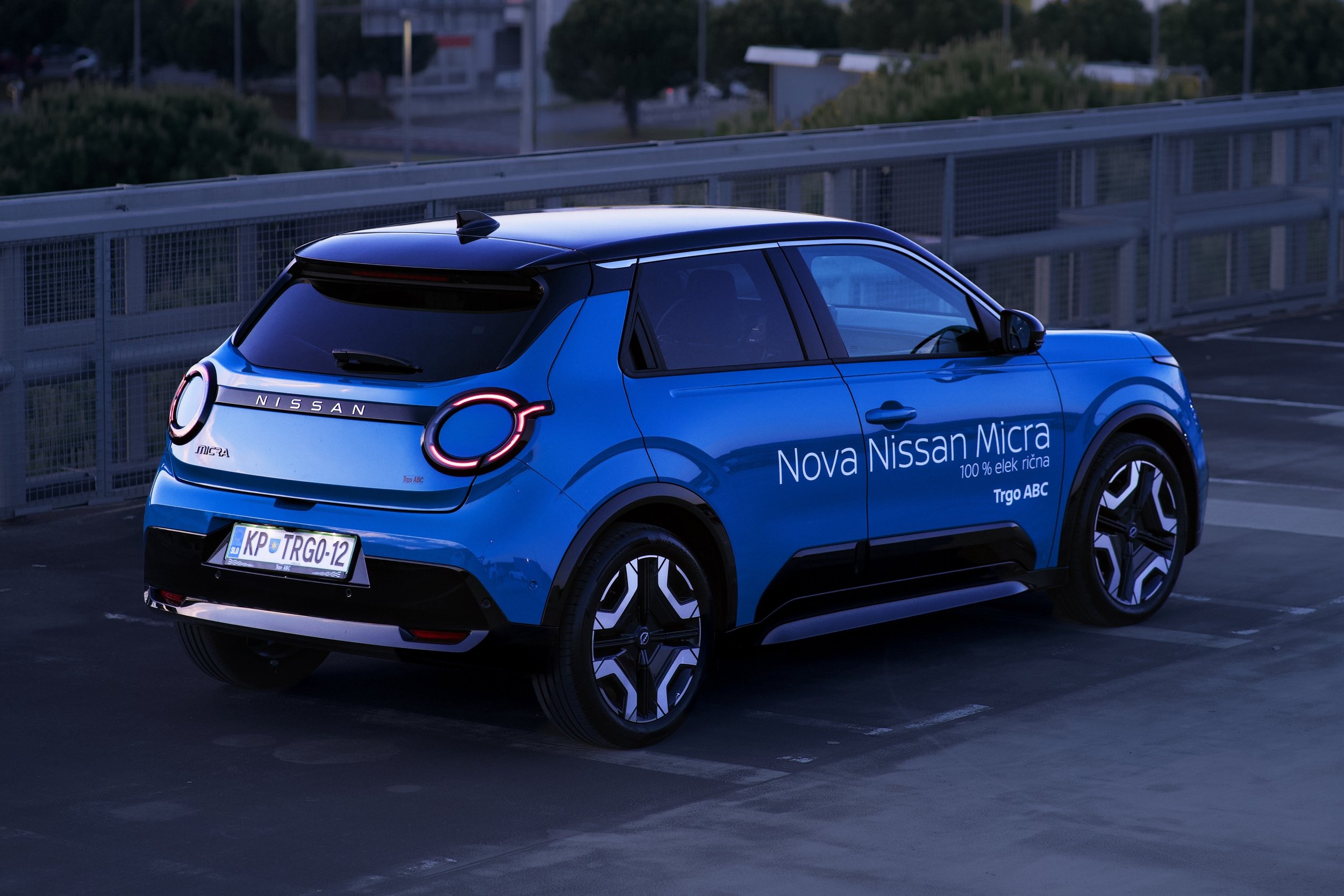 Blue Nissan Micra compact car parked in an outdoor parking lot at dusk, with visible text indicating it is a 100% electric vehicle.
