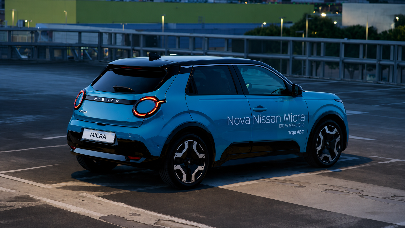 Blue Nissan Micra parked in an outdoor multi-level parking garage at dusk. The car has illuminated taillights and the side of the car displays text promoting it as a 100% electric vehicle.