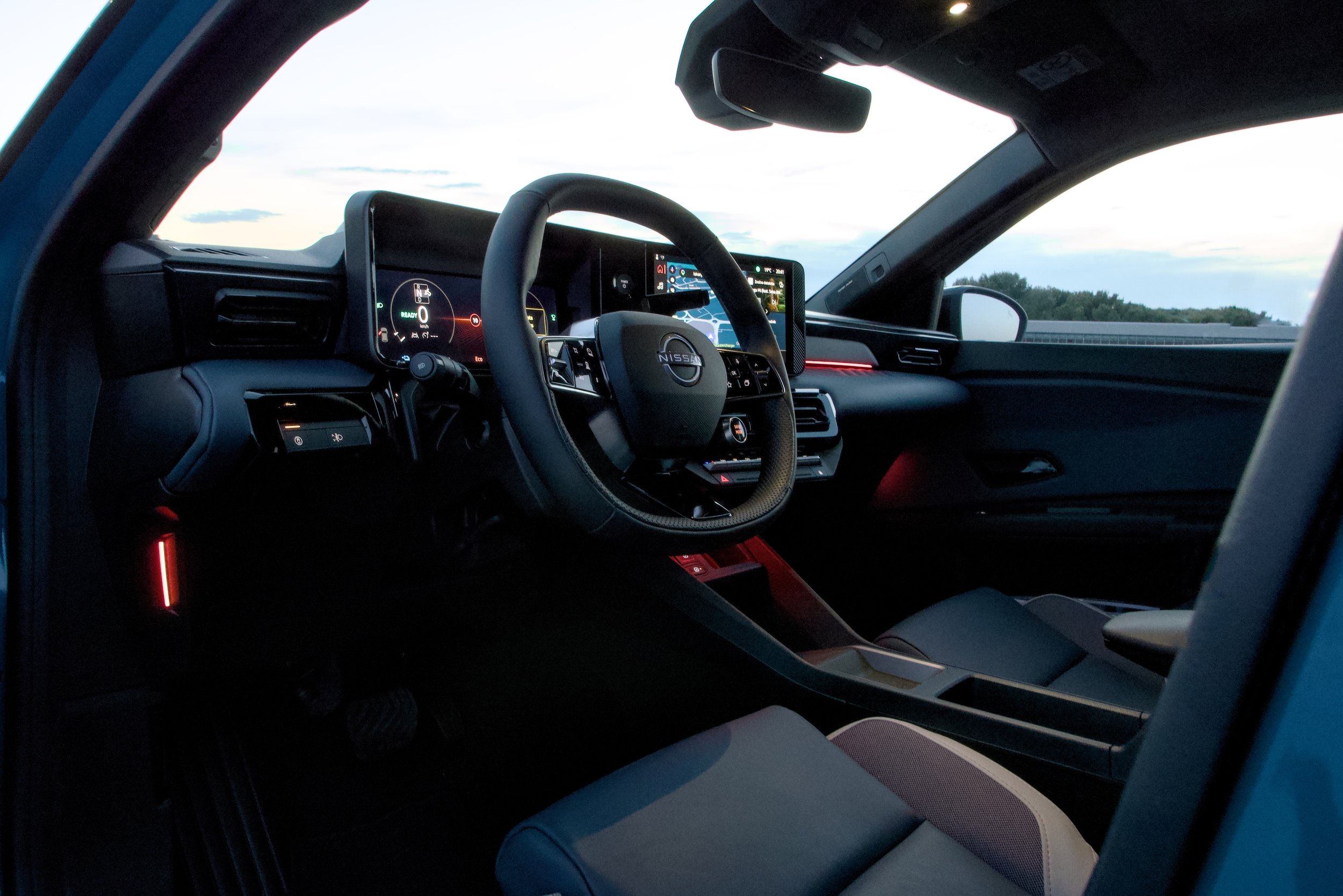 Interior of a Nissan vehicle, showing the steering wheel, dashboard with digital displays, and a near view of the front passenger seat, during twilight with a view of the outdoors through the windshield.
