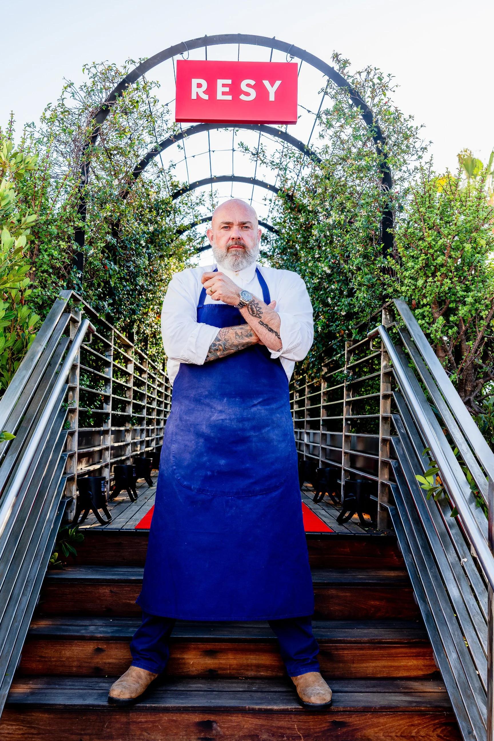 Chef Evan Fluke with a beard and tattoos wearing a white shirt and blue apron, standing on stairs with metal railings, in front of greenery, with a red sign that says 'RESY' hanging above him.