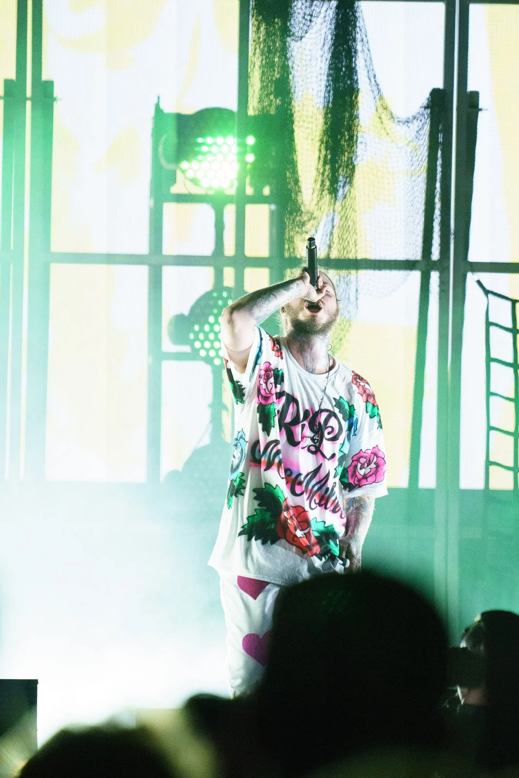 Post Malone singing into microphone on stage with colorful lighting and background visuals.