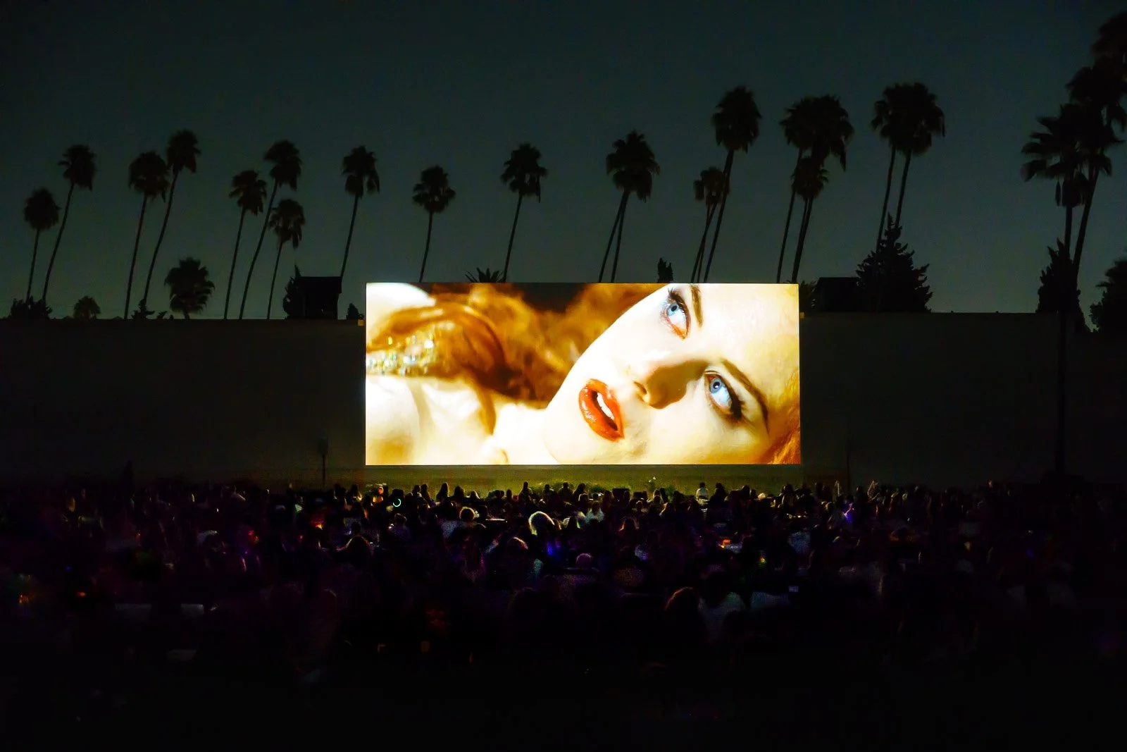 Outdoor movie screening at night with a large screen displaying a close-up of a woman's face with blue eyes and red hair, surrounded by a crowd of seated viewers and tall palm trees silhouetted against the dark sky.