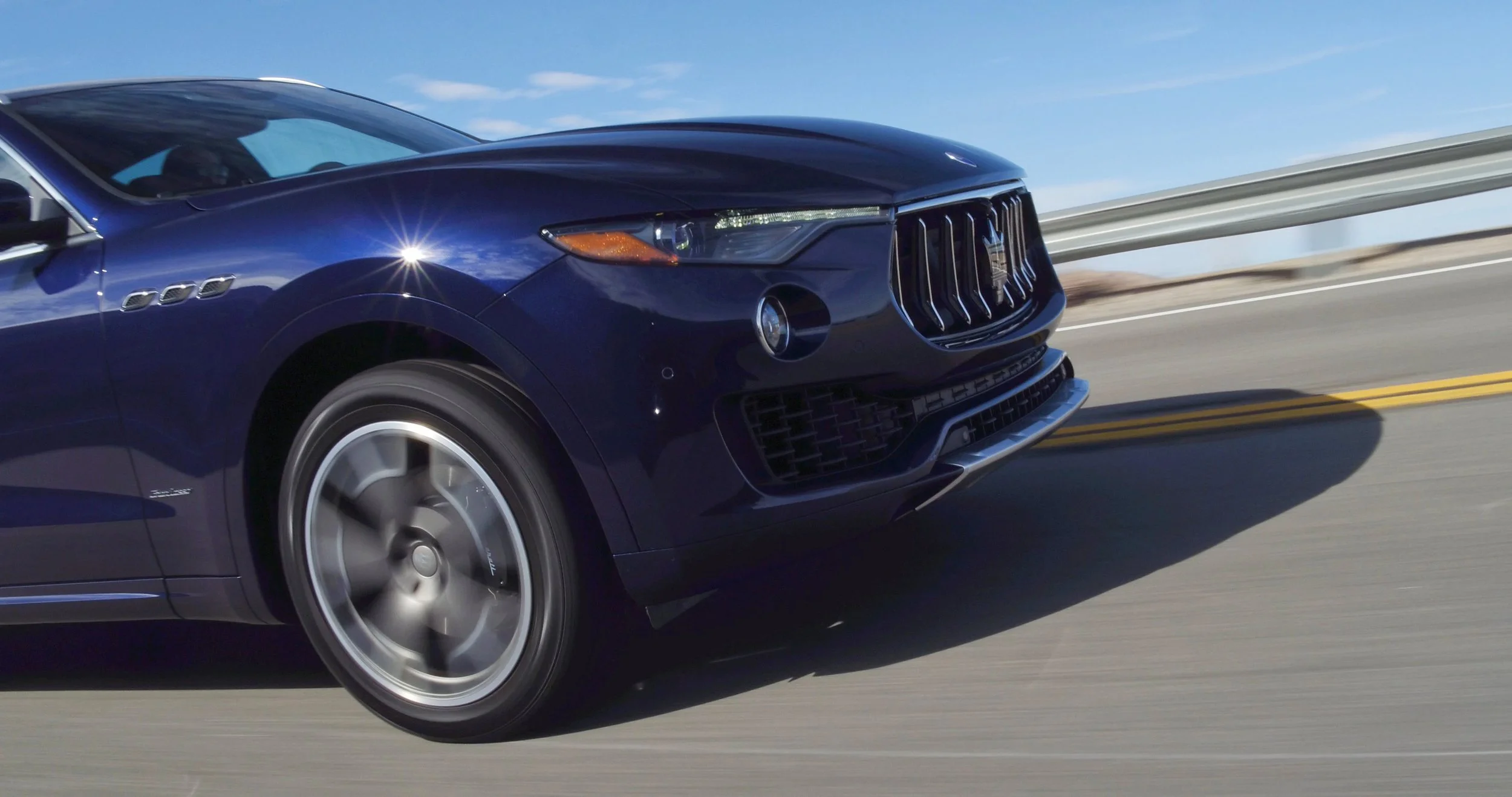 Maserati Levante - a dark blue luxury SUV driving on an open highway with a metal guardrail on the side and a clear blue sky in the background.