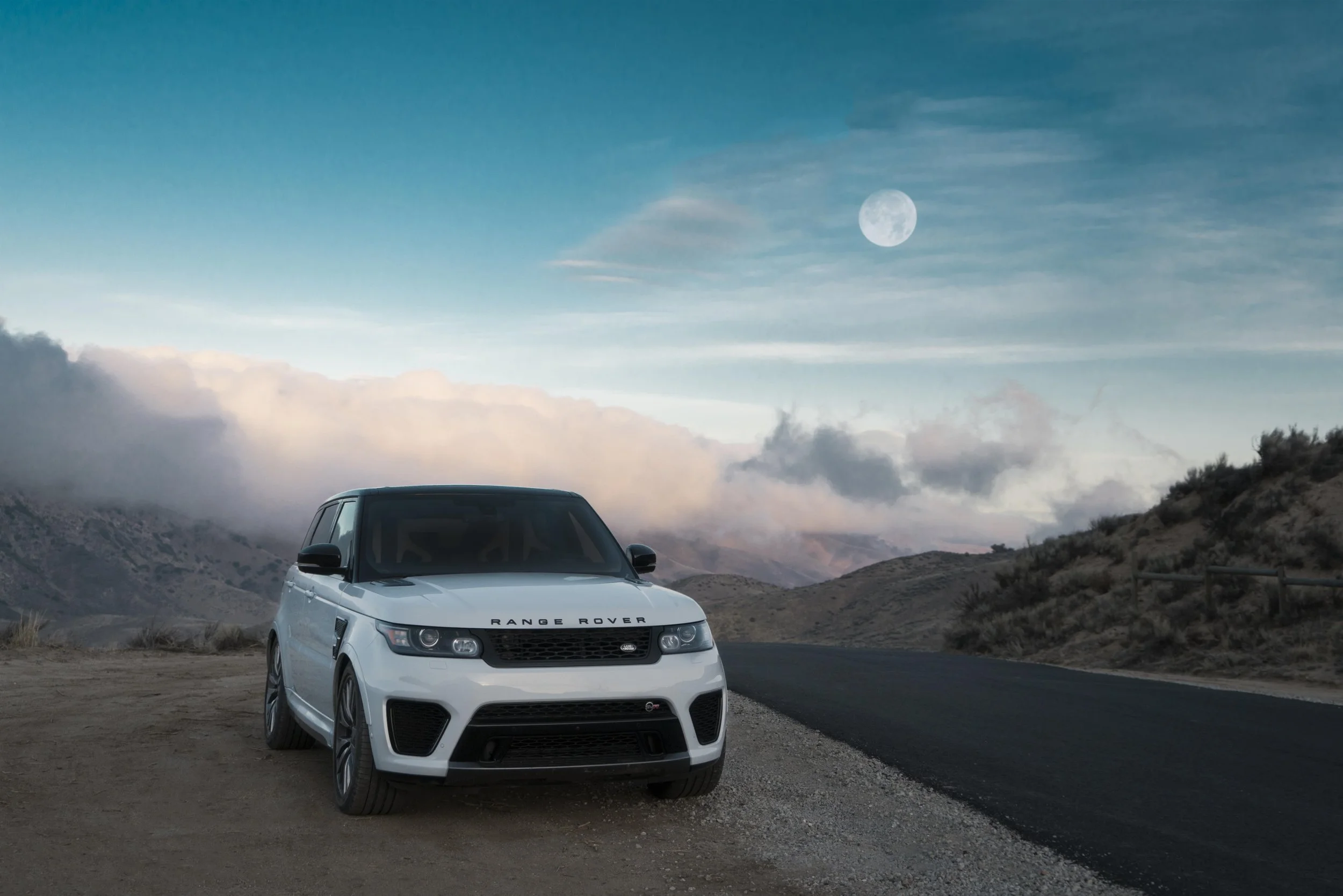A white Range Rover parked on the side of a winding mountain road at dusk, with a full moon in the sky and cloudy weather above.