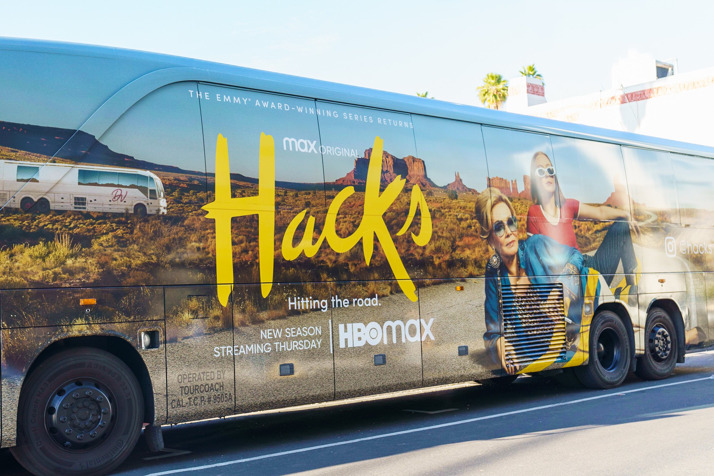 A bus with a desert landscape and mountain scenery, advertising a new season of the TV series 'Hacks' on HBO Max, featuring two women in sunglasses.